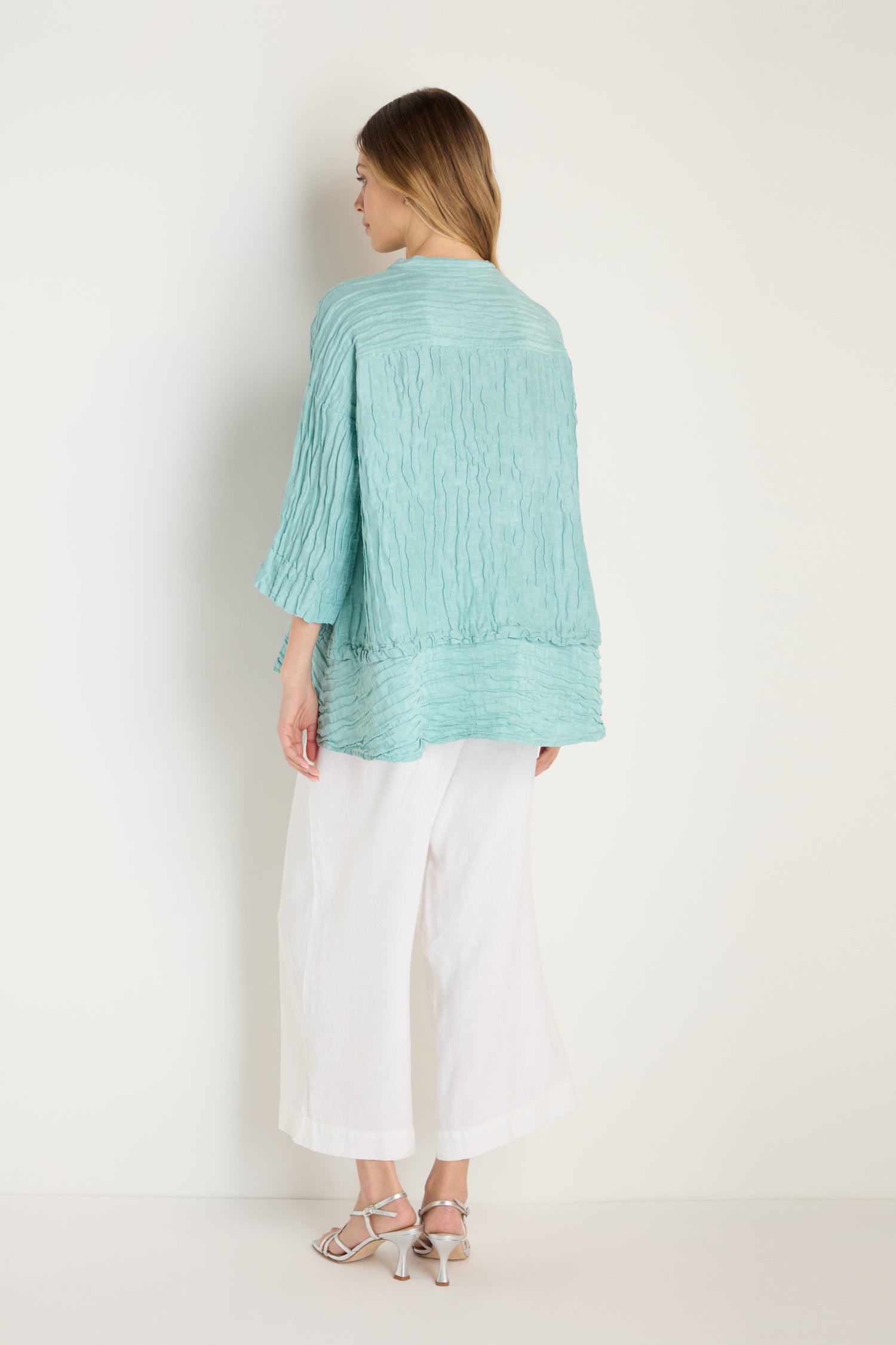 A woman faces away against a white wall, wearing the turquoise Panel Hem Silk Linen Shirt with an oversized, breathable fit, paired with wide white pants and white strappy sandals.