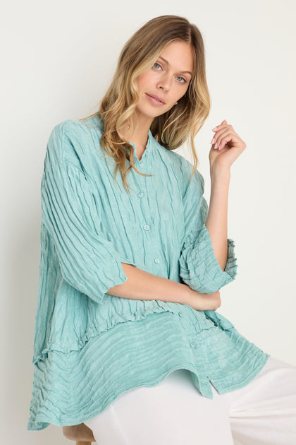Woman in the Panel Hem Silk Linen Shirt, featuring a loose, breathable fit with buttons and an oversized style, paired with white pants against a plain light background.