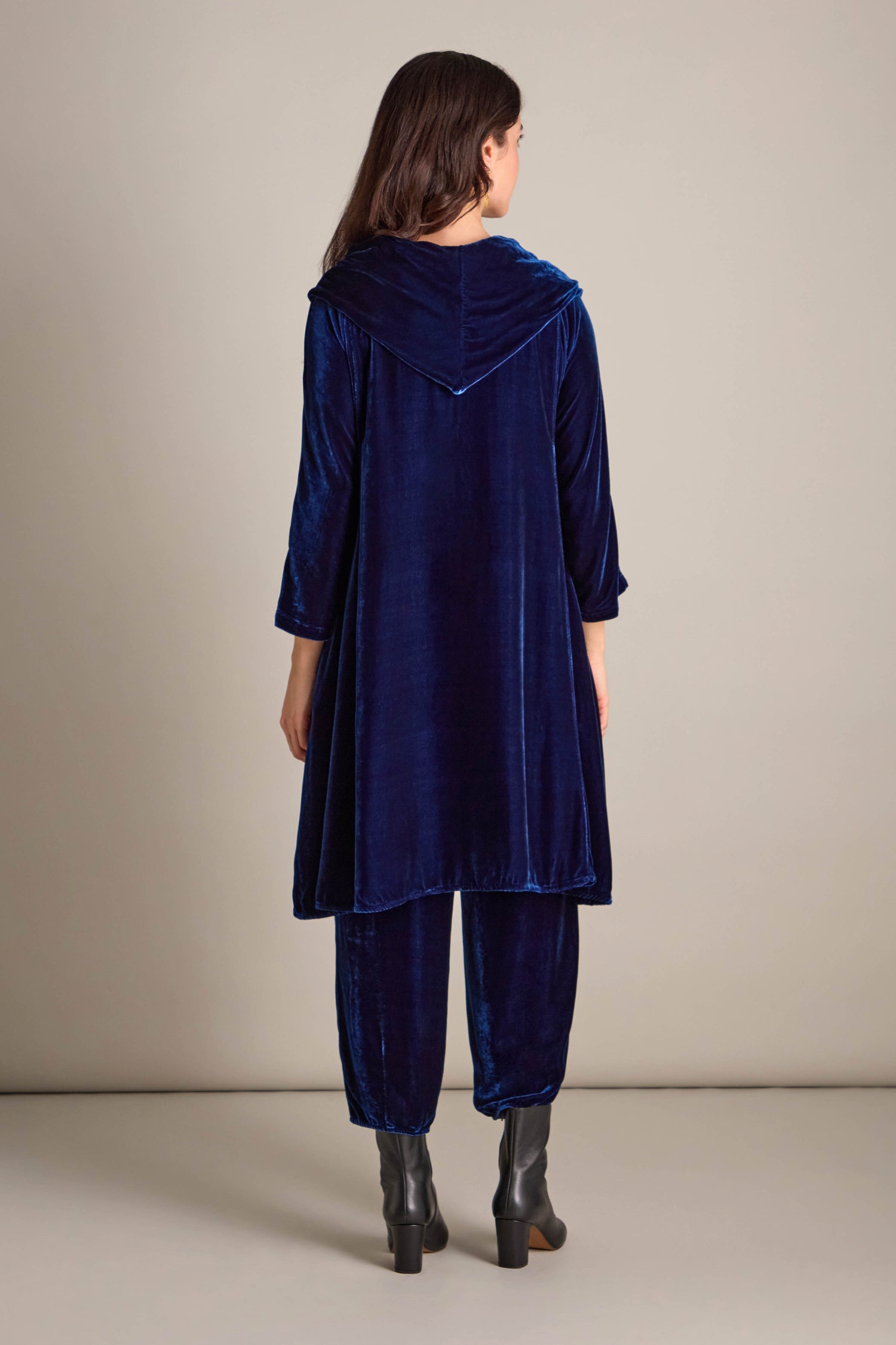 A person in a stunning Cowl Silk Velvet Dress, featuring a rich dark blue hue with a hooded tunic and pants, stands facing away. The ensemble is perfectly paired with sleek black ankle boots.