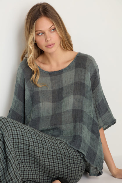 A woman with long, wavy hair sits against a white wall, wearing the Large Check Boxy Top and matching plaid pants in muted green and gray tones, both made from soft woven linen with a large check pattern.