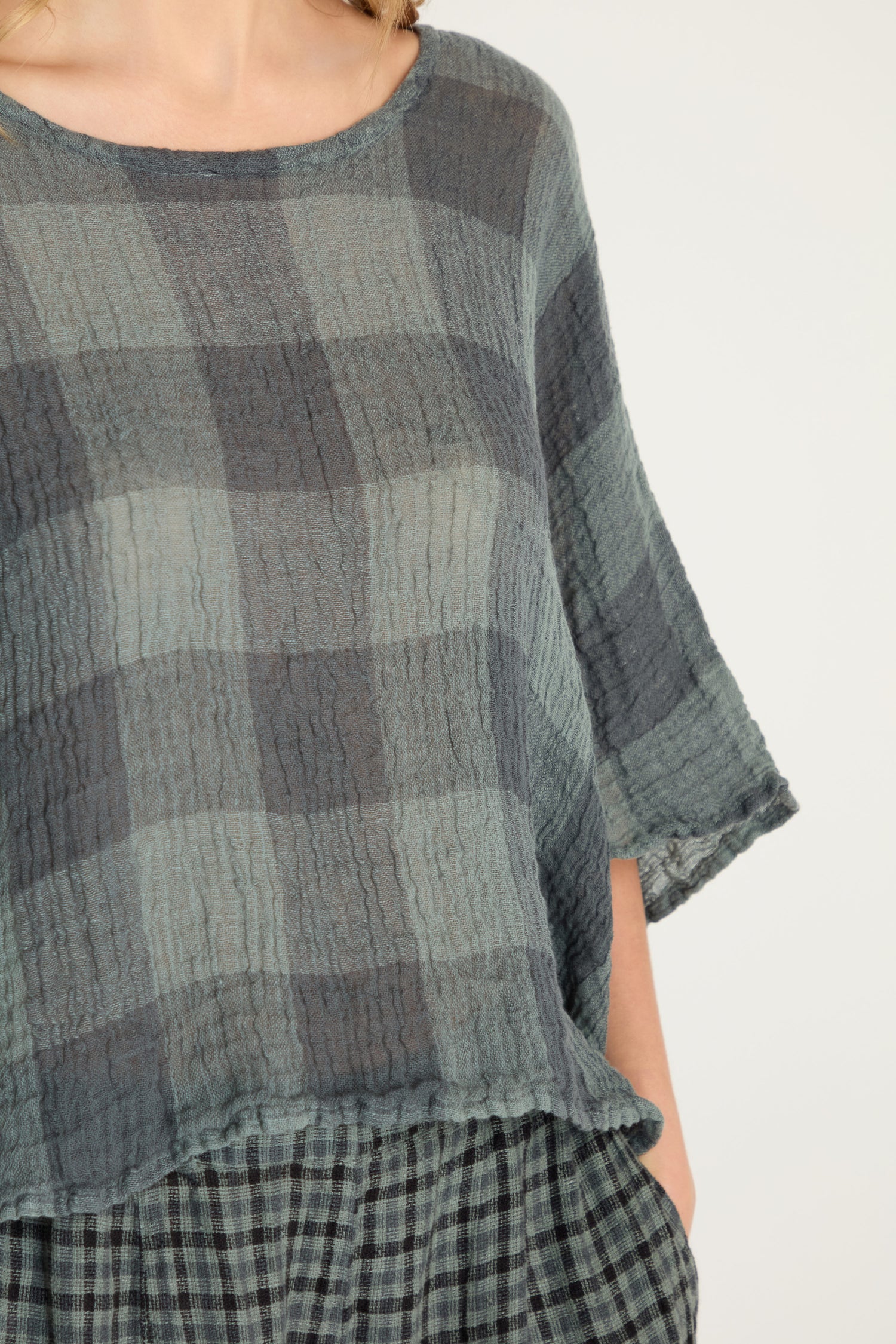 Model wearing the Large Check Boxy Top, a loose-fitting, boxy top in a gray and blue textured large check woven linen. Image shows the upper torso and part of the arm.