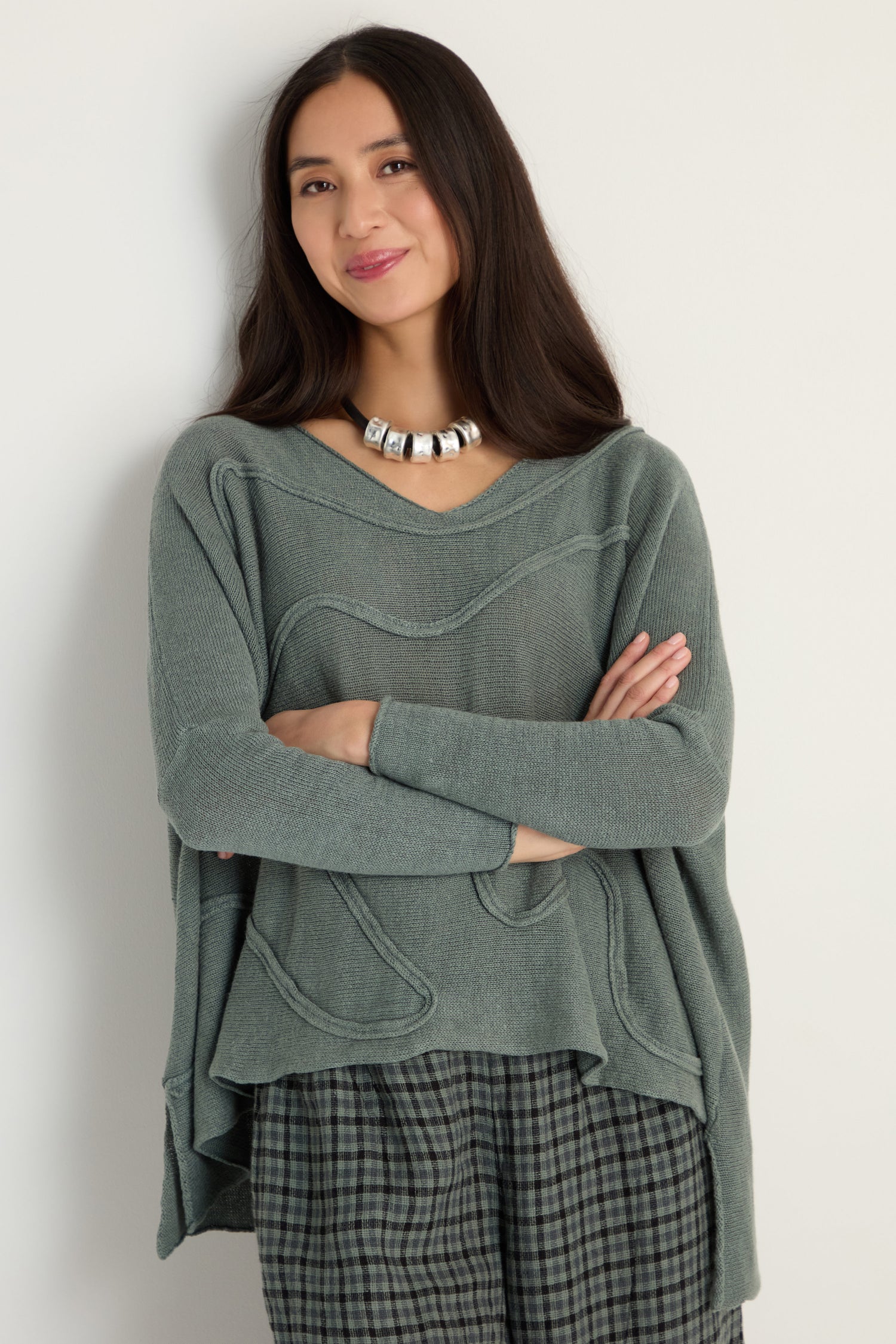 Woman with long dark hair wears the Swirl Seamed Linen Knit Top in green, paired with a silver necklace and plaid pants, standing against a plain light background with arms crossed.