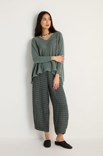 A woman stands against a white wall wearing the Swirl Seamed Linen Knit Top in green, styled with checkered wide-leg pants, a beaded necklace, and black slip-on shoes.