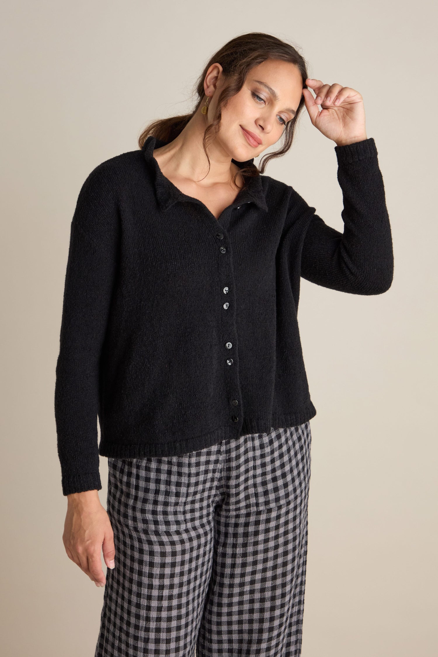 A person wearing a Frill Neck Wool Cardi in black and checkered pants, touching their hair with one hand, stands against a plain backdrop, showcasing relaxed styling.