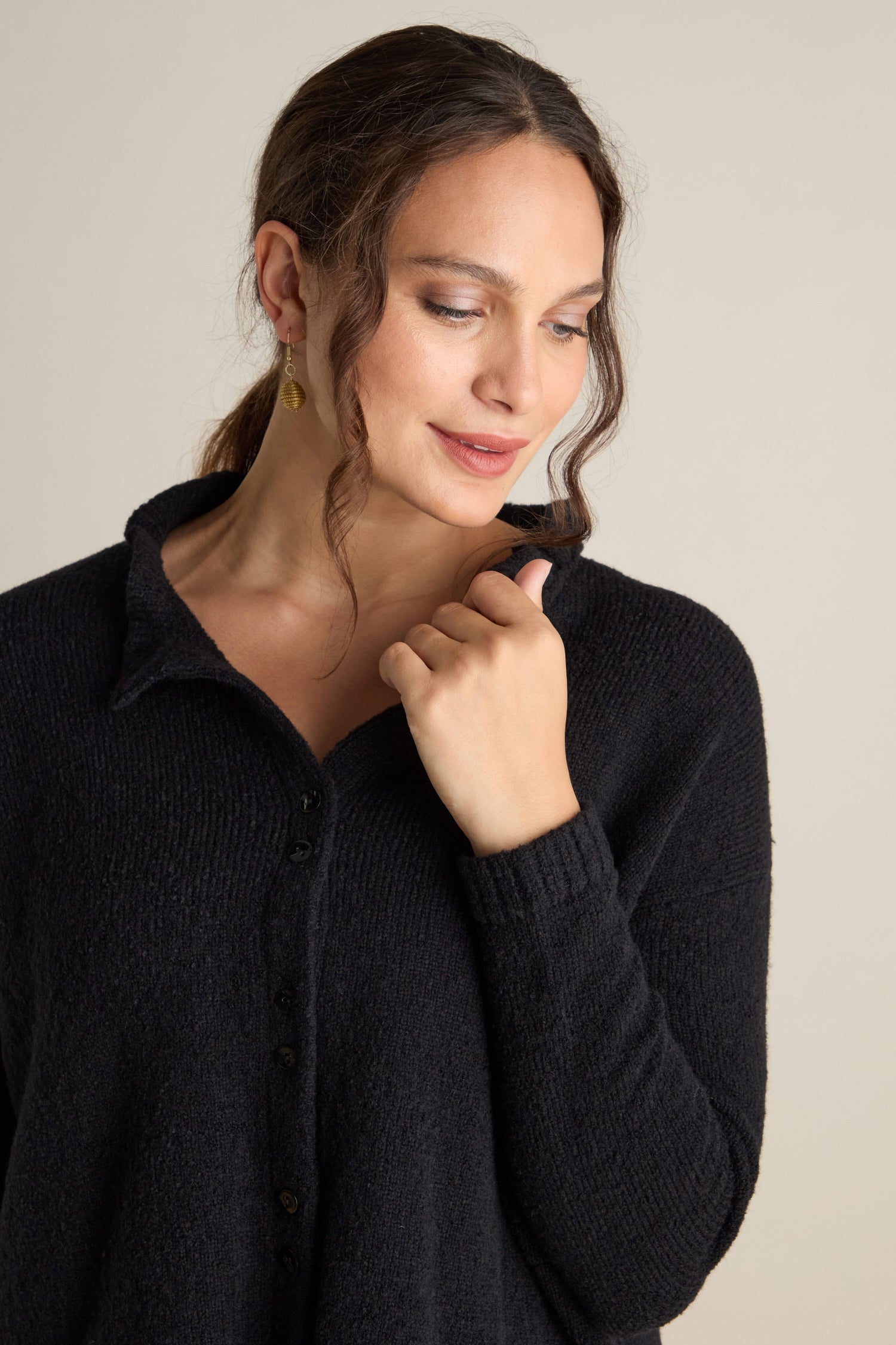 A woman with wavy hair tied back is wearing a dark-colored Frill Neck Wool Cardi, looking down with a slight smile on her face.