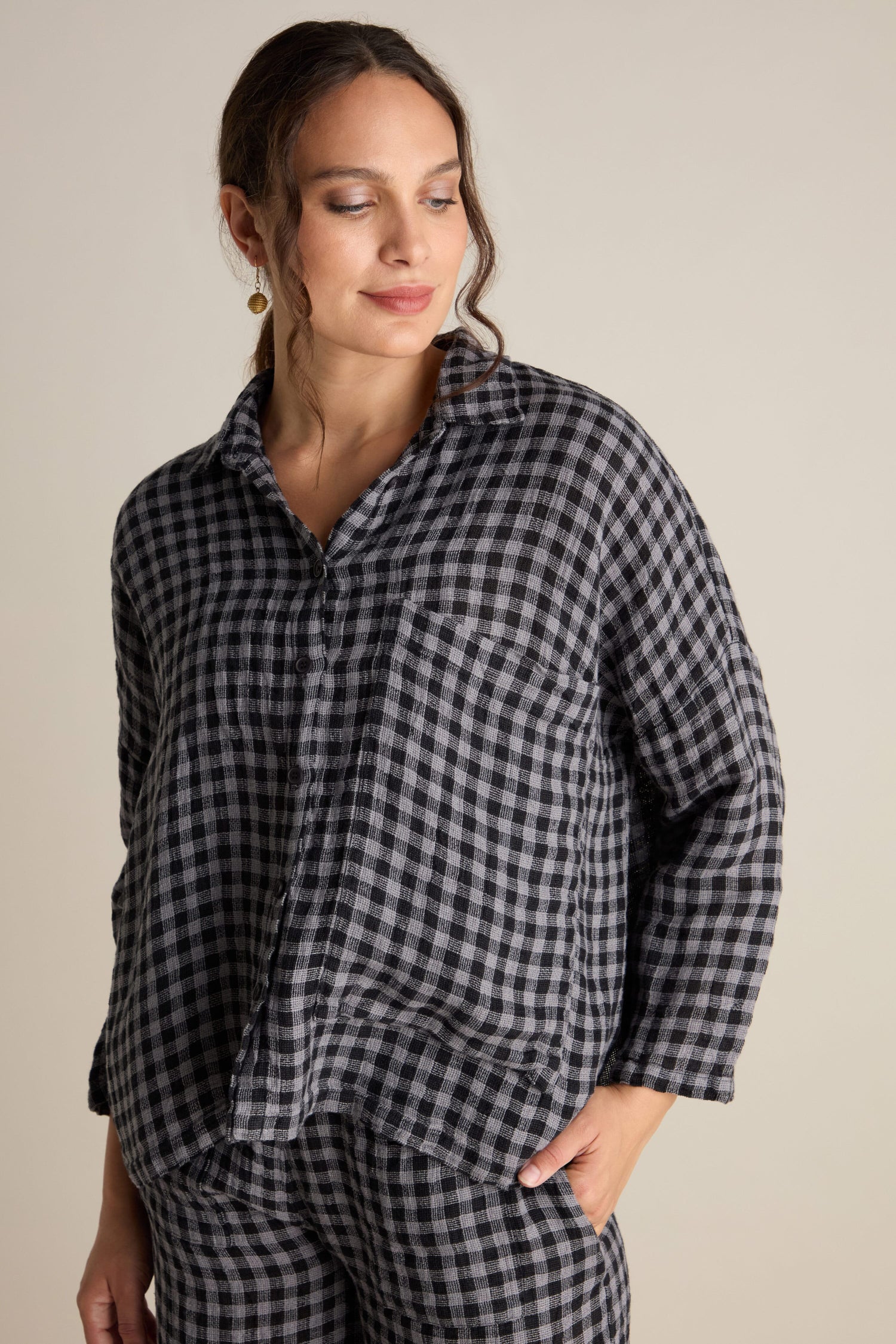 A person wearing the Linen Check Big Pocket Shirt, a black and white checkered long-sleeve linen shirt with an oversized pocket, along with matching pants, stands against a plain background, perfect for day-dressing.