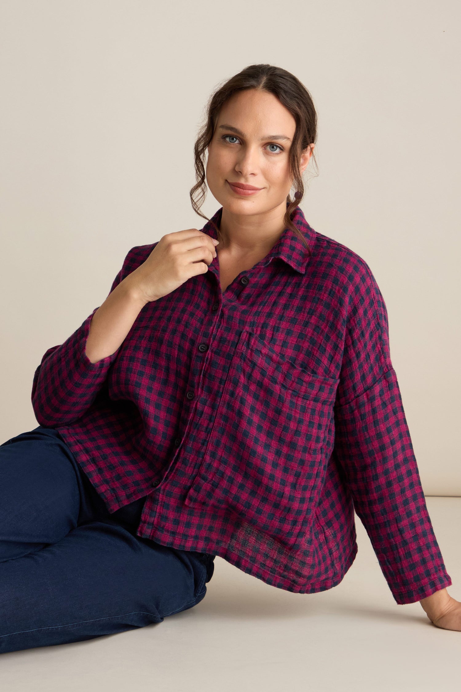 A person with dark hair is seated against a beige background, wearing the Linen Check Big Pocket Shirt in red and black plaid, paired with dark blue pants.