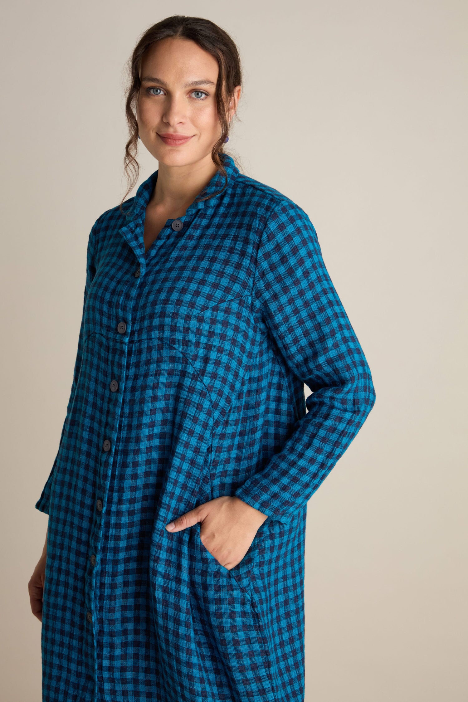 A person in a Linen Check Shirt Dress stands against a plain beige background, with one hand in their pocket and the other arm relaxed by their side, subtly contrasting the loose-fitting style of breathable linen.