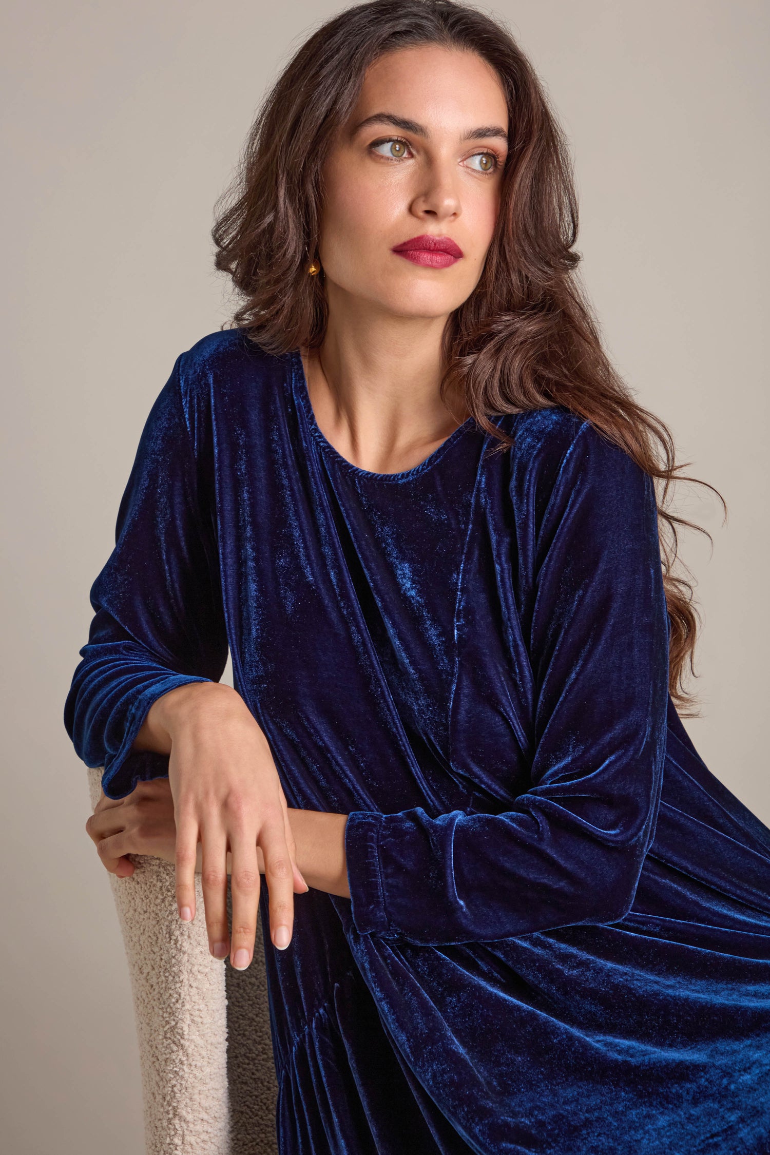 A woman in the Drape Front Silk Velvet Dress, characterized by its asymmetric design, sits gazing to the side with a neutral expression, embodying luxury and elegance.