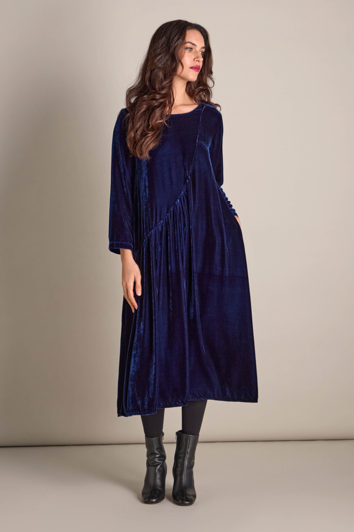 Dressed in the luxurious Drape Front Silk Velvet Dress, a woman stands against a plain background. Her long brown hair cascades down gracefully, enhancing her sophisticated appearance, while she completes her elegant ensemble with black ankle boots.