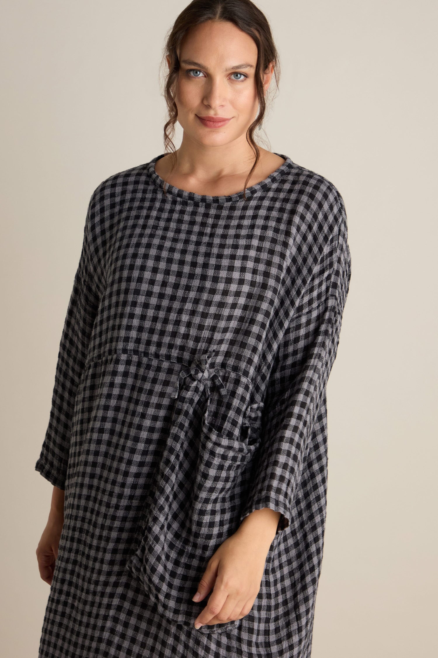 A person with wavy hair is wearing a loose-fitting, long-sleeved Linen Check Big Pocket Dress featuring an oversized pocket, standing against a plain backdrop.