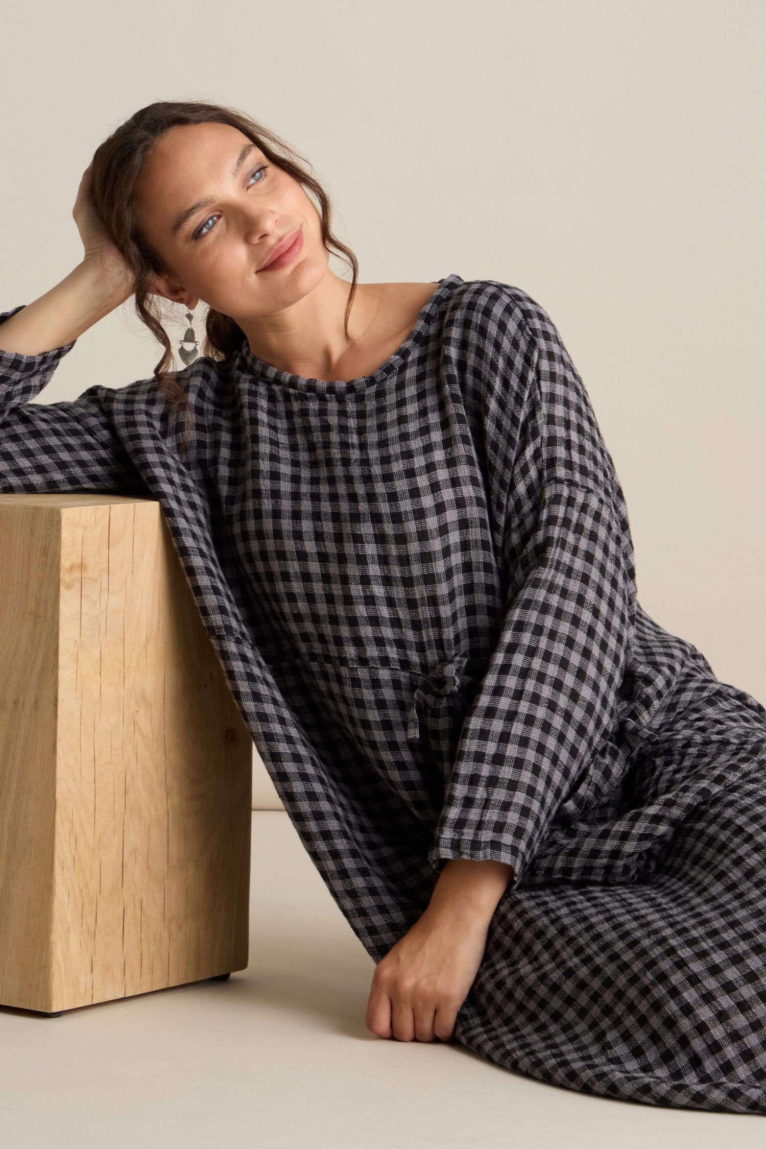 A woman in the Linen Check Big Pocket Dress leans against a wooden surface with a relaxed expression, effortlessly showcasing its oversized feature pocket.