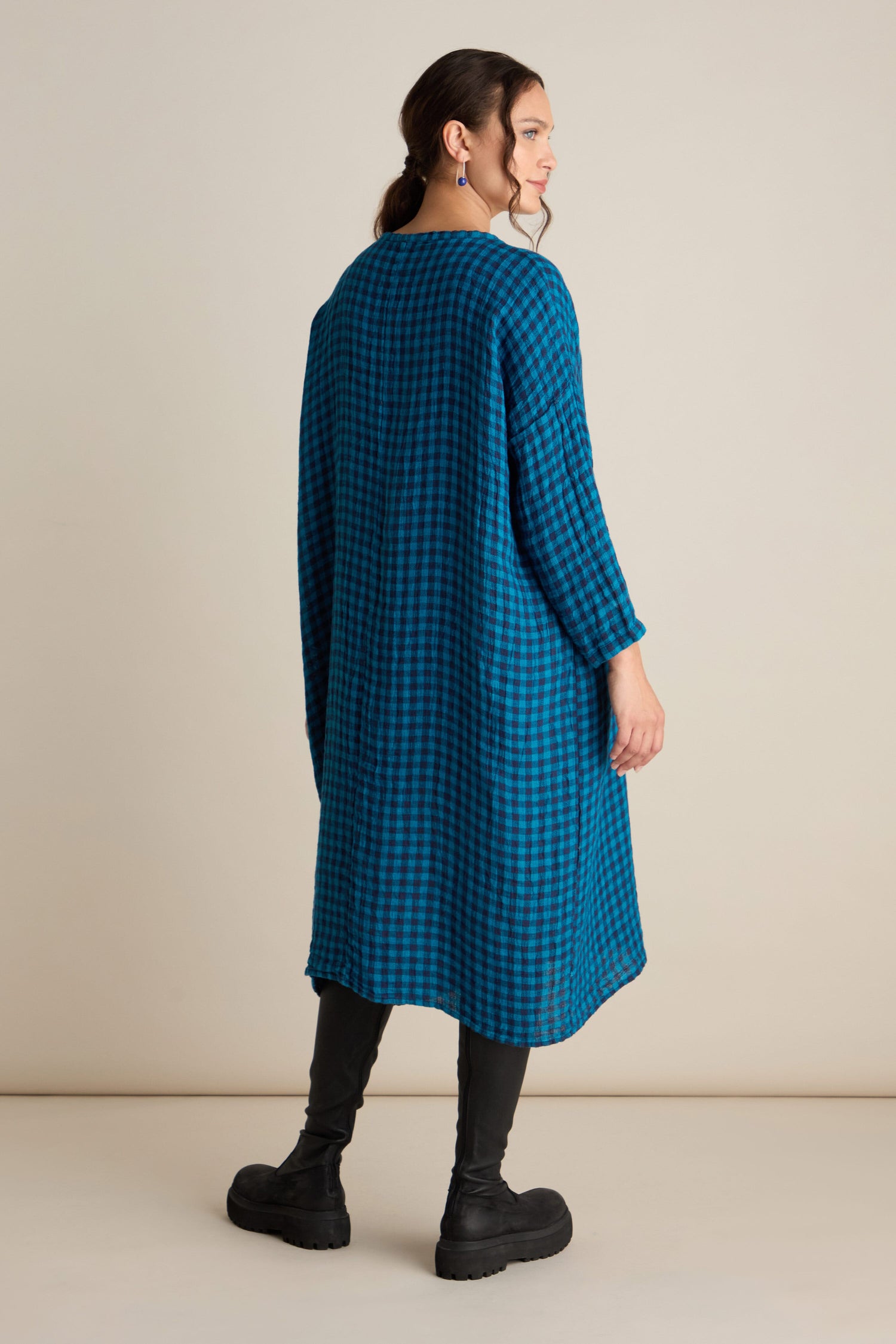 A person stands against a plain background, wearing the Linen Check Big Pocket Dress—an oversized, long-sleeved, knee-length dress made from soft linen with a blue check pattern. They pair it with black leggings and black shoes. The relaxed tunic style of the dress adds comfort while its large feature pocket enhances its casual charm.