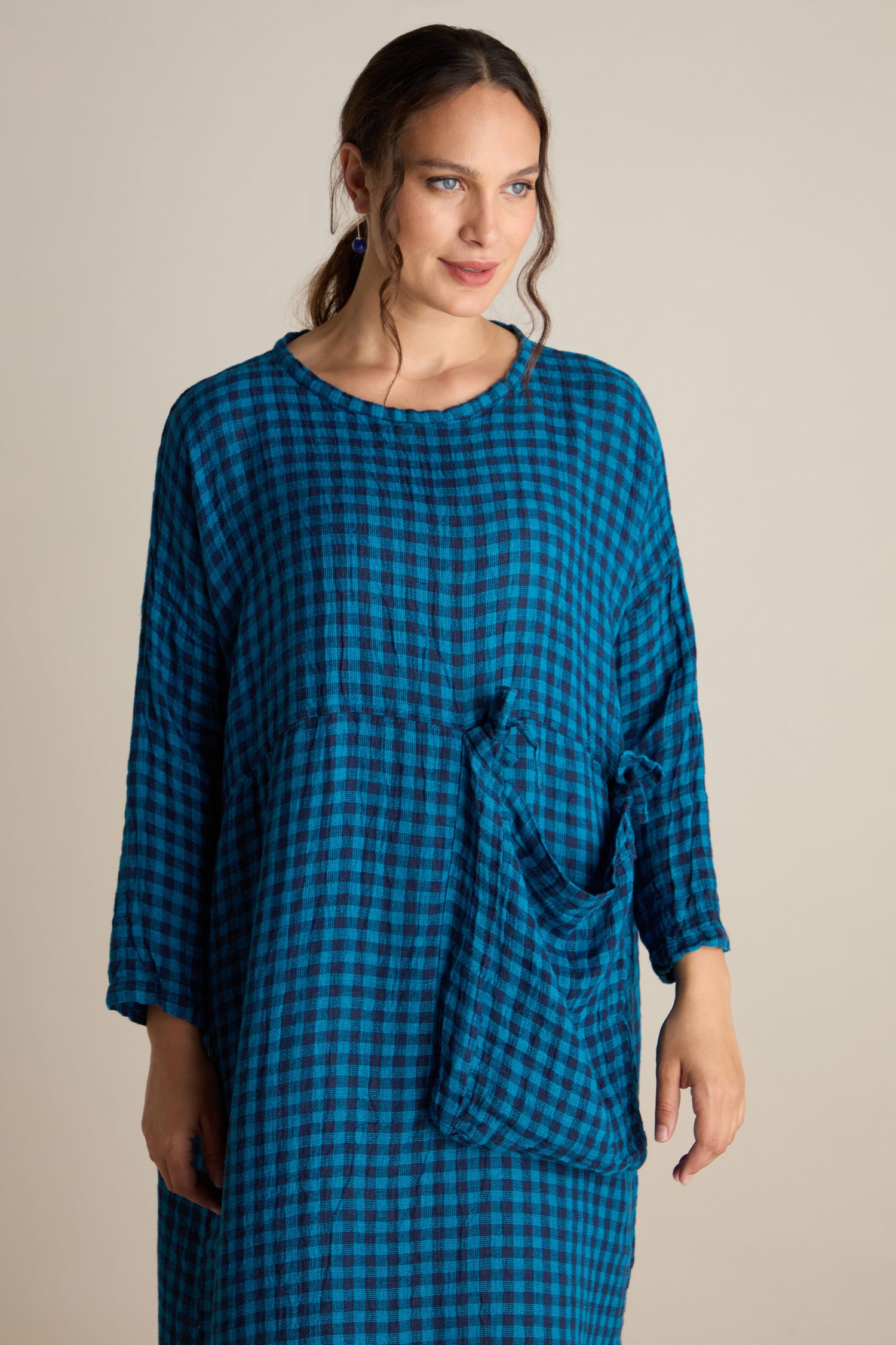 A woman wearing the Linen Check Big Pocket Dress, which is a loose-fitting blue and black checkered dress featuring an oversized pocket, stands against a neutral background.