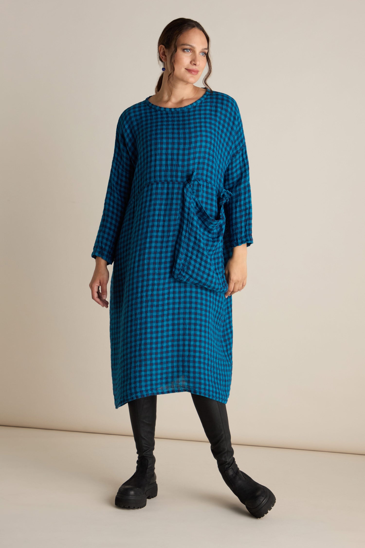 A woman stands wearing the Linen Check Big Pocket Dress, showcasing its blue, checkered design with long sleeves and a relaxed tunic style featuring an oversized pocket. She pairs it with black boots and faces slightly to the side against a beige background.