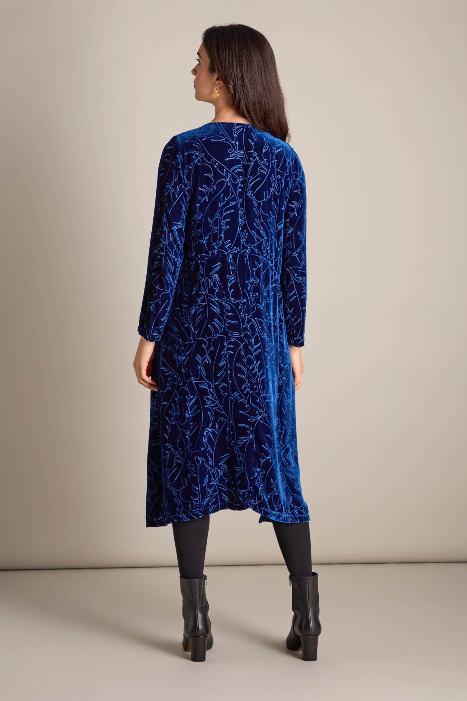 A woman with long hair is wearing a blue patterned Vine Devore Dress and black boots, standing with her back to the camera against a neutral background, showcasing an ideal piece for day-to-evening wear.