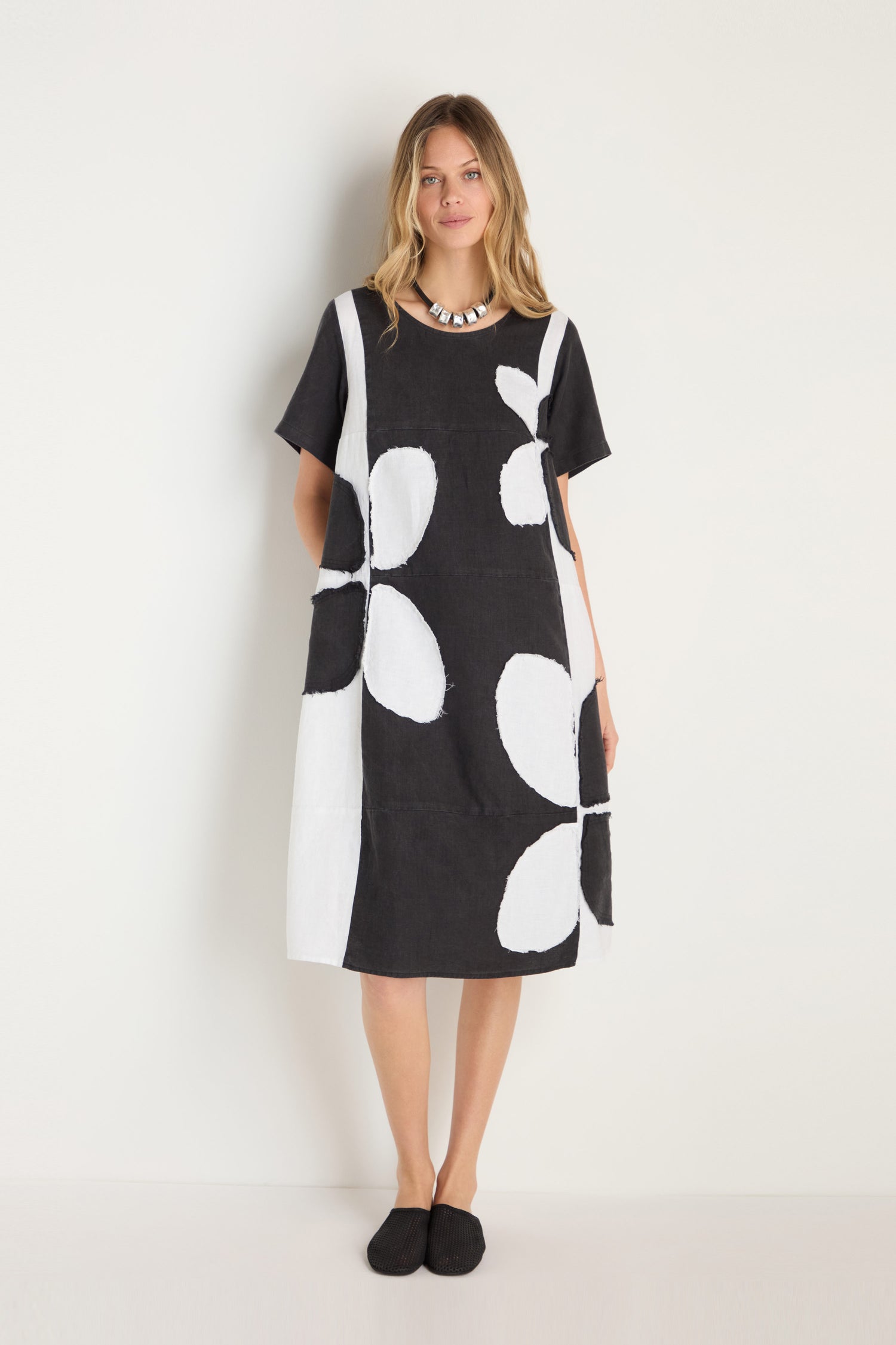 A woman stands against a white wall wearing the Quant Monochrome Linen Dress with large white floral patterns, paired with a chunky necklace and black shoes.