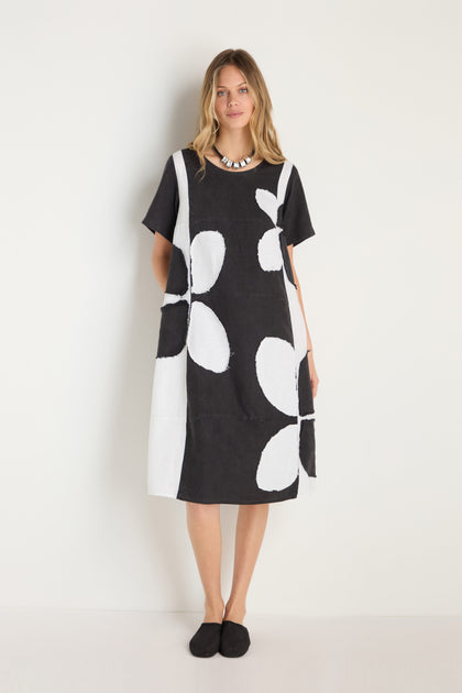 A woman stands against a white wall wearing the Quant Monochrome Linen Dress with large white floral patterns, paired with a chunky necklace and black shoes.