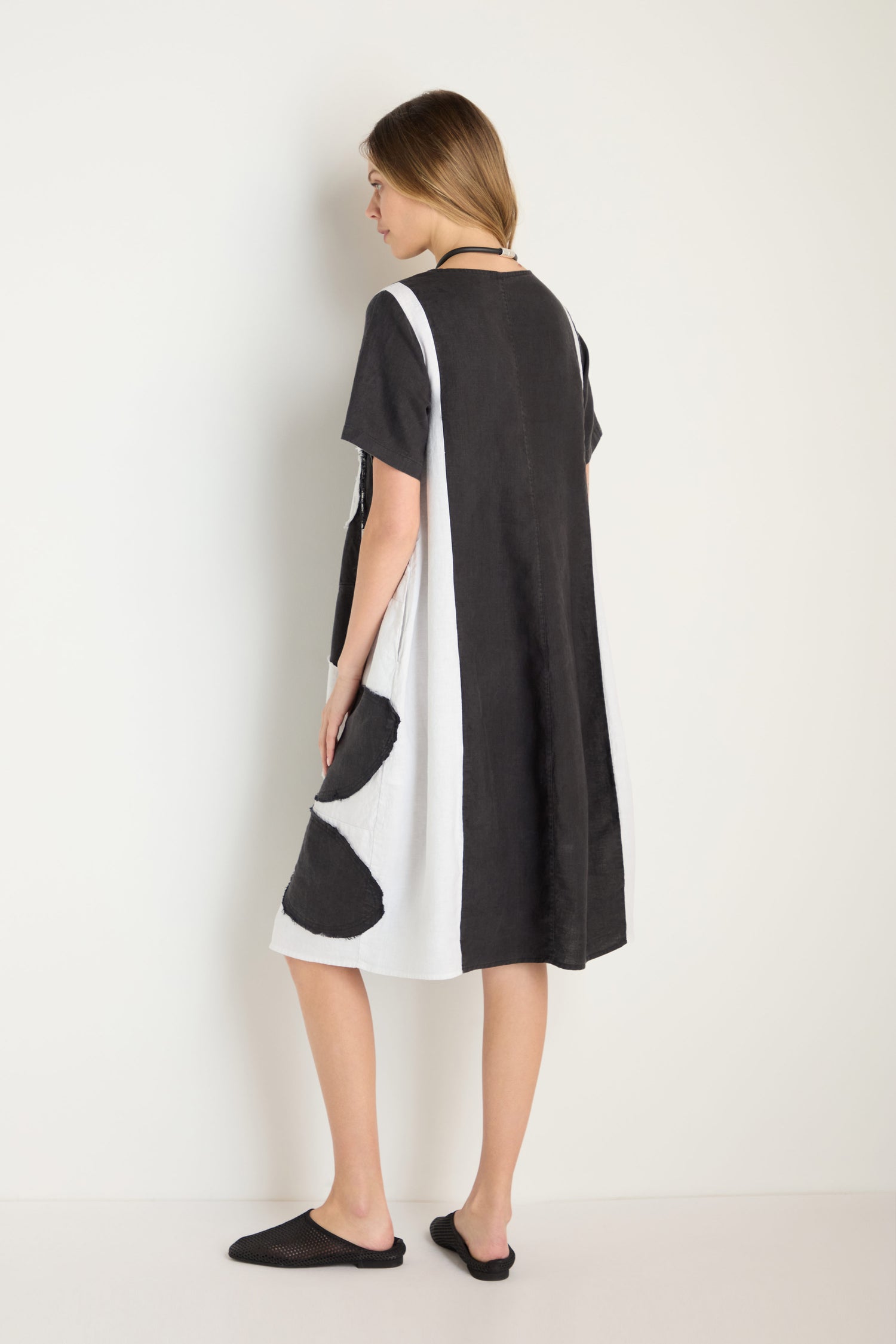 A woman faces away, wearing the Quant Monochrome Linen Dress with black and white color-blocking and black slip-on shoes, set against a plain light background.