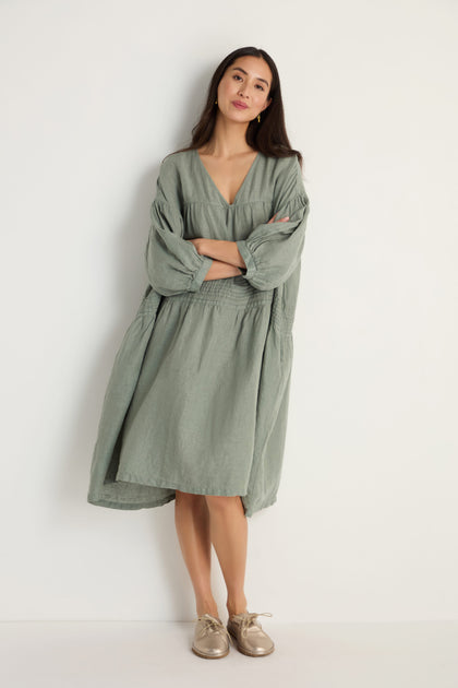 A woman with a neutral expression stands against a white wall, arms crossed, wearing the Easy Fit V-Neck Sage Linen Dress and beige shoes.