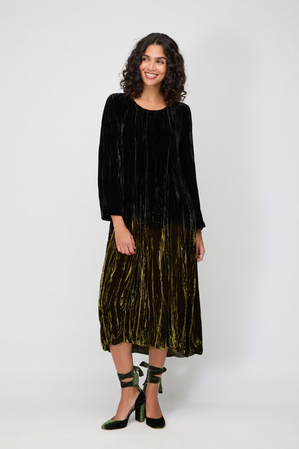 A woman stands against a plain background, wearing the Silk Velvet Ombre Dress—a long-sleeve black velvet top flowing into an olive-green skirt—paired with green ankle-tie heels.