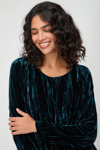 A woman with dark, curly hair smiles with her eyes closed, wearing the Crushed Velvet Peacock Dress, standing against a plain light background.