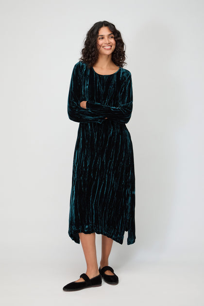 A woman with curly hair, smiling with arms lightly crossed, wears the Crushed Velvet Peacock Dress—long-sleeved and dark green—paired with black slip-on shoes against a plain light background.