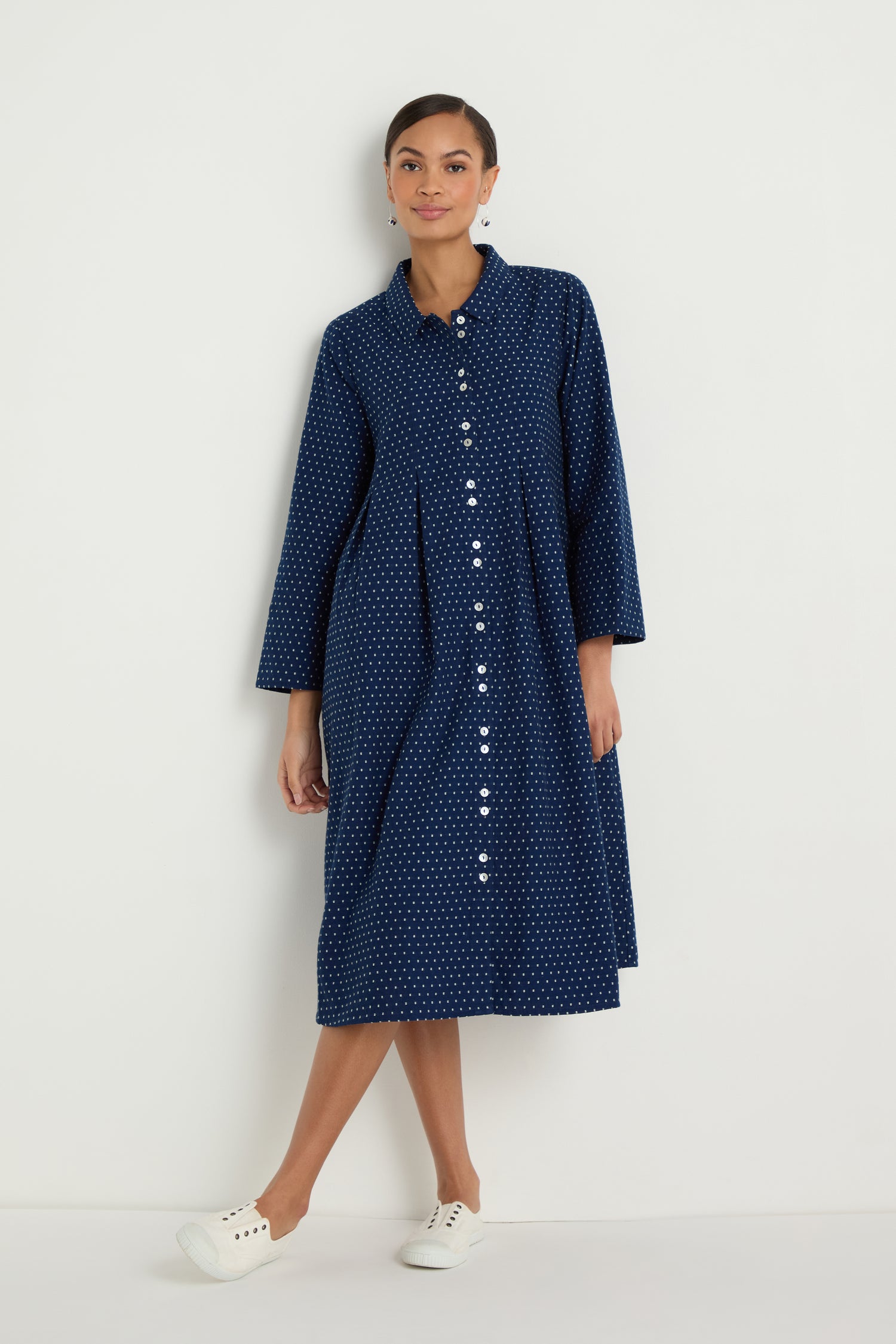 A person wears the Indigo Spot Cotton Shirt Dress—long-sleeved, navy blue with small white polka dots and an A-line silhouette—paired with white sneakers, standing against a plain white background.