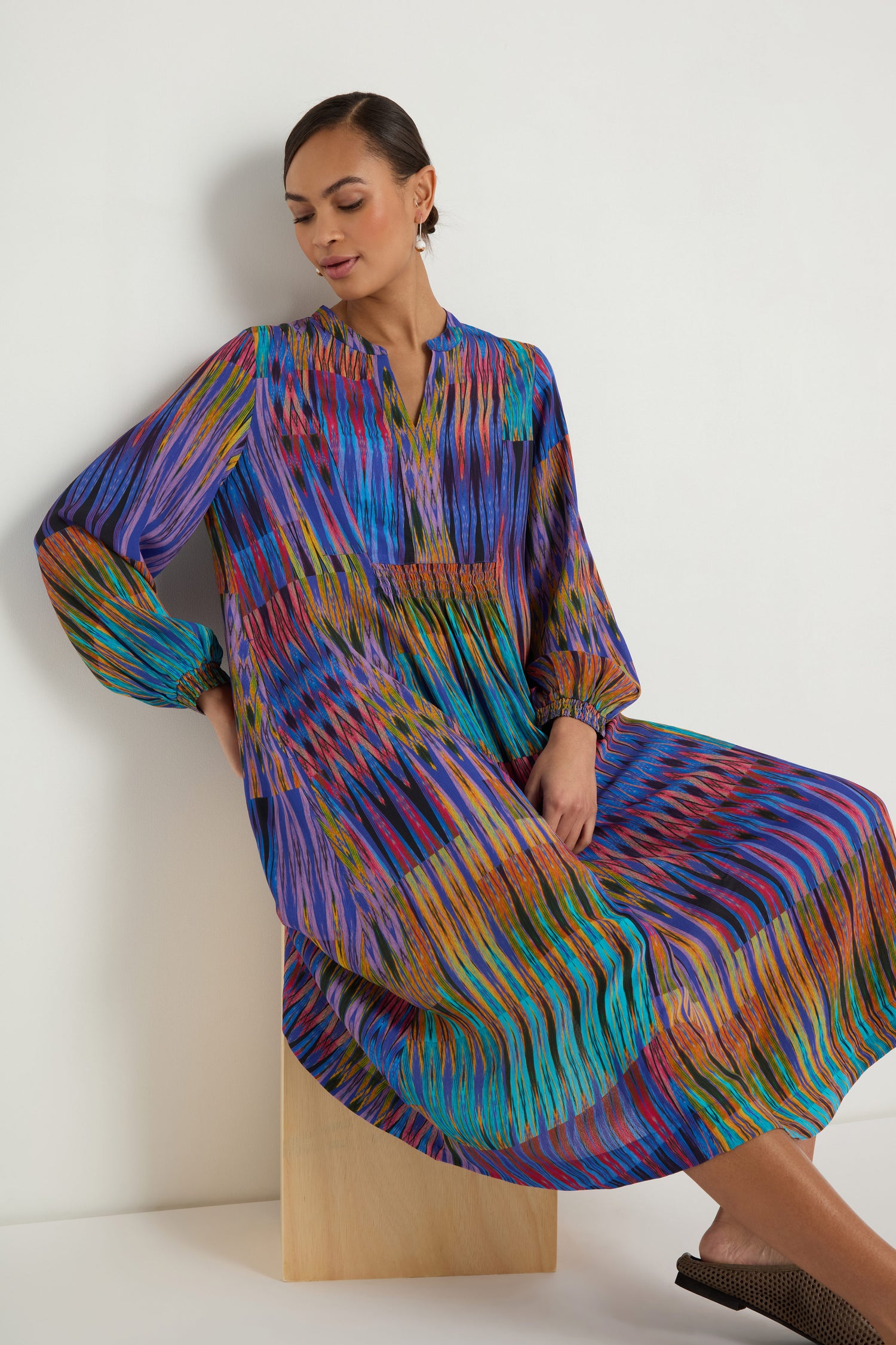 A woman sits on a wooden block, wearing the Vivid Ikat Patchwork Dress, which features a relaxed fit, long sleeves, and a mid-length hem.