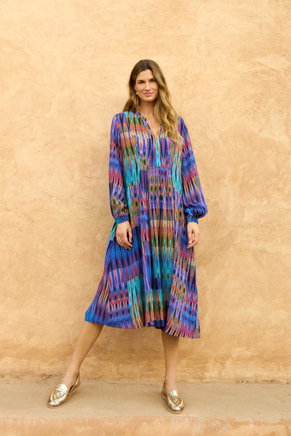A woman stands against a textured beige wall wearing the Vivid Ikat Patchwork Dress, a long-sleeved, midi-length piece, paired with gold flat shoes.