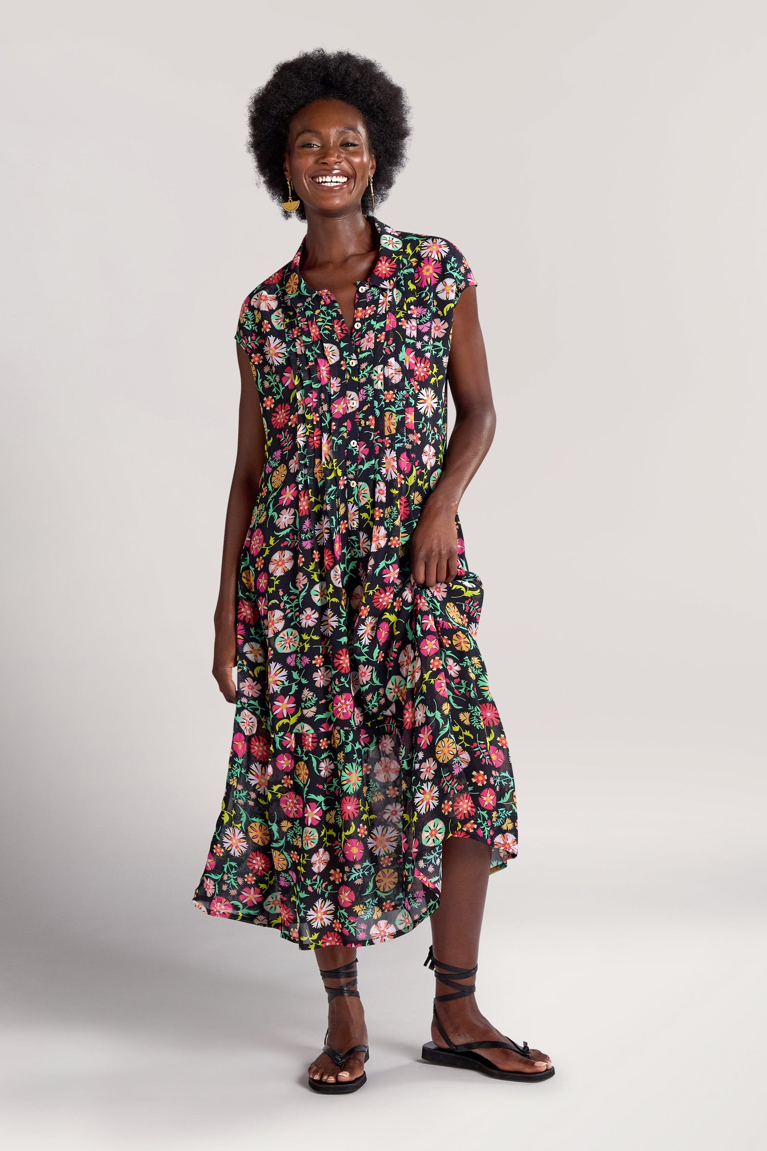 A person wearing a sleeveless, Folkloric Floral Shirt Dress stands with one leg slightly forward and an arm resting by their side while smiling. They have short curly hair and wear sandals.