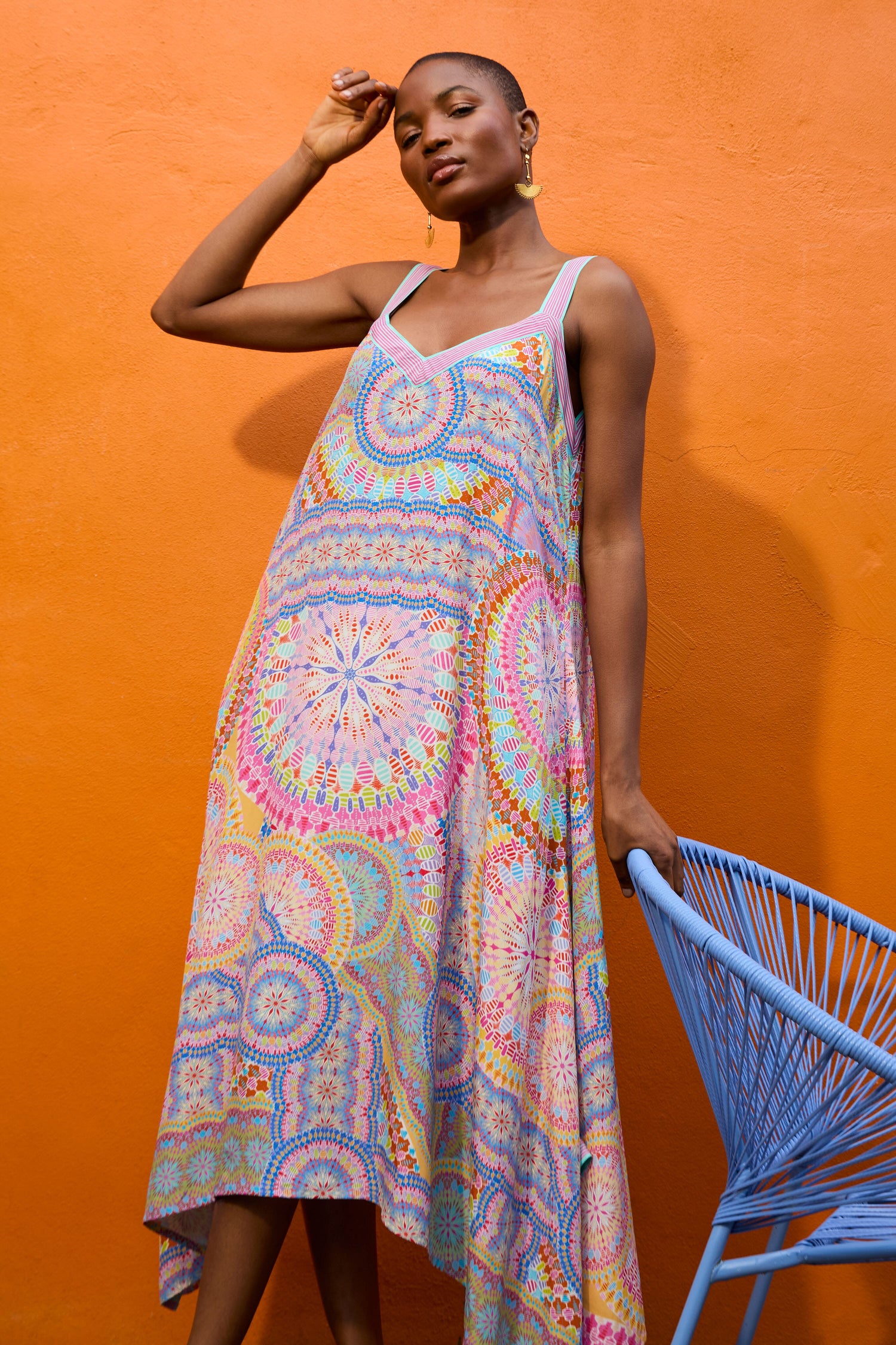 A woman wearing a colorful Circle Mandala Sleeveless Dress stands against an orange wall, gently touching her head with one hand while the other rests on a blue chair.