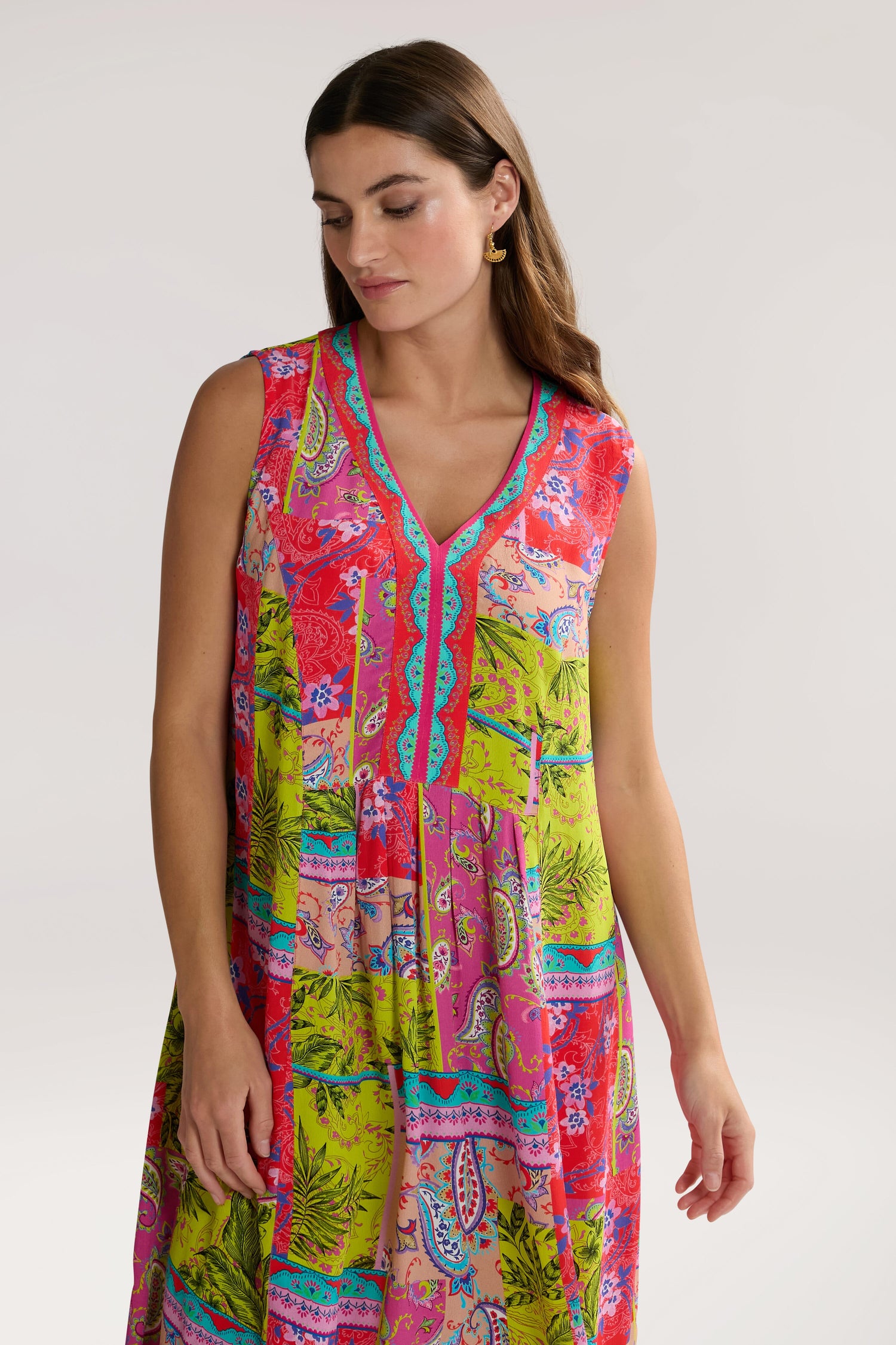 A woman with long brown hair is wearing a Tropical Patchwork Sleeveless Bubble Dress featuring a mix of pink, green, and red patterns. She is looking down with a neutral expression, embodying a relaxed bohemian style.