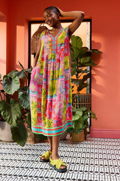 A person stands smiling in a vibrant room with large plants and a coral-colored wall, wearing a Tropical Patchwork Sleeveless Bubble Dress and yellow sandals, with one hand touching their head.