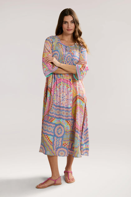 A woman stands with arms crossed, wearing a colorful Circle Mandala Pleat Front Dress and pink sandals against a plain background.