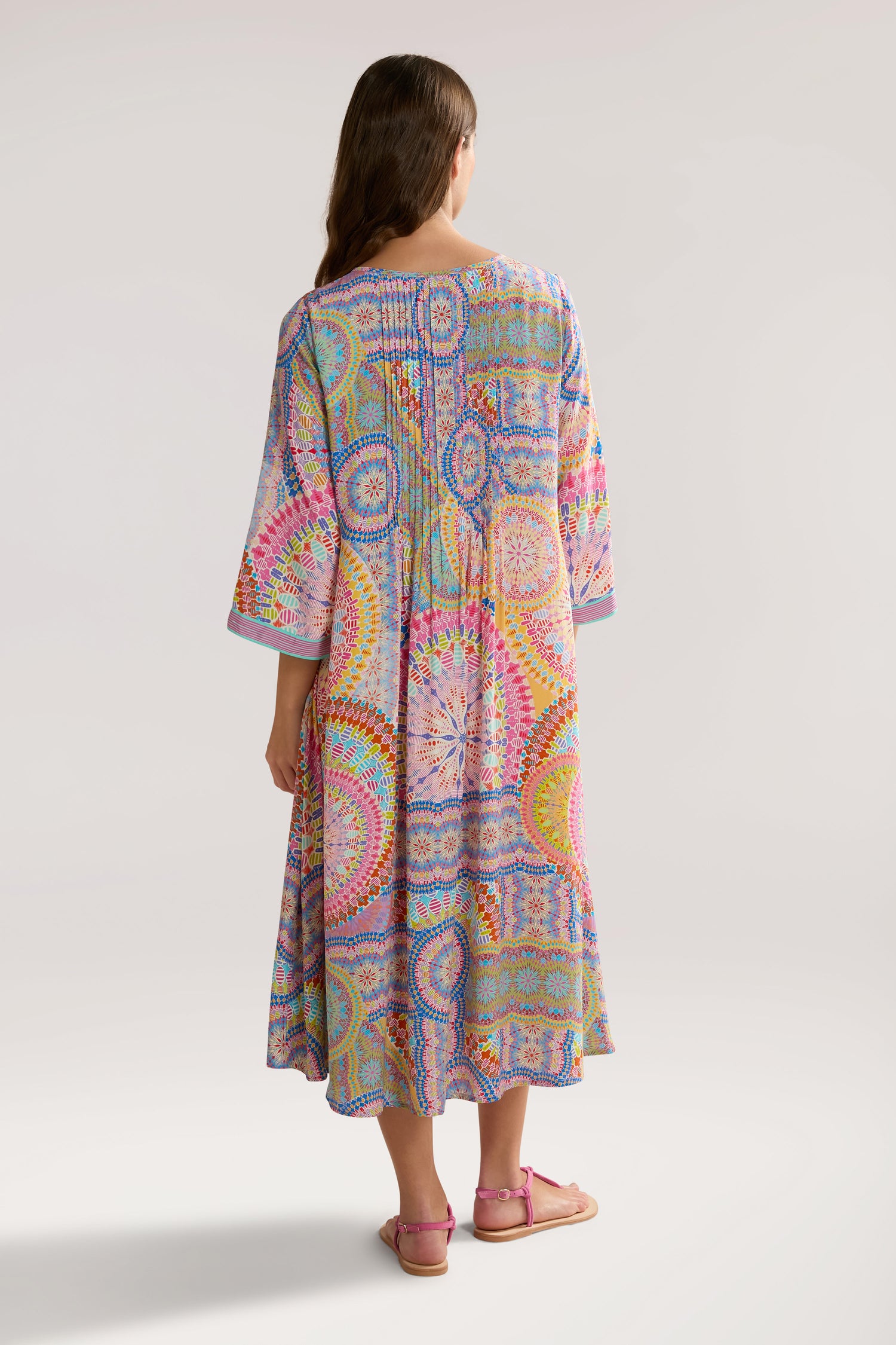 A woman stands facing away, wearing a colorful, patterned Circle Mandala Pleat Front Dress with three-quarter sleeves. She has long hair and is wearing pink sandals.