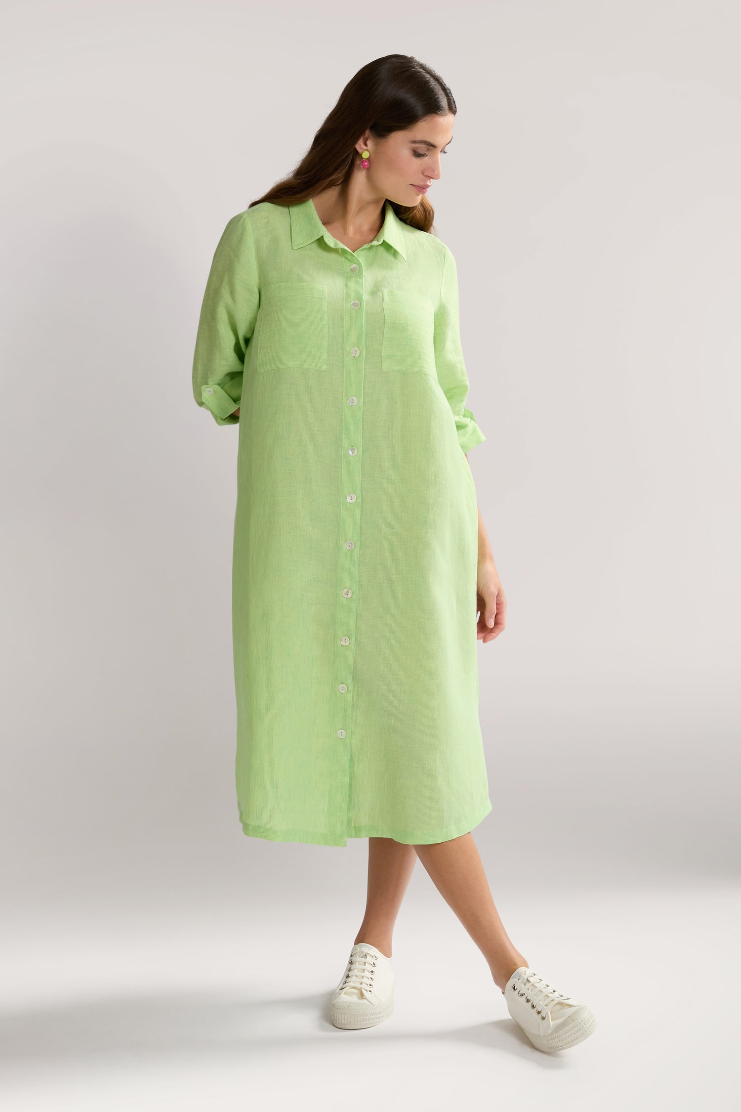 A woman is wearing a light green, button-down, long-sleeve Linen Ticking Stripe Shirt Dress paired with white sneakers. She is standing against a plain background and looking down to her left.