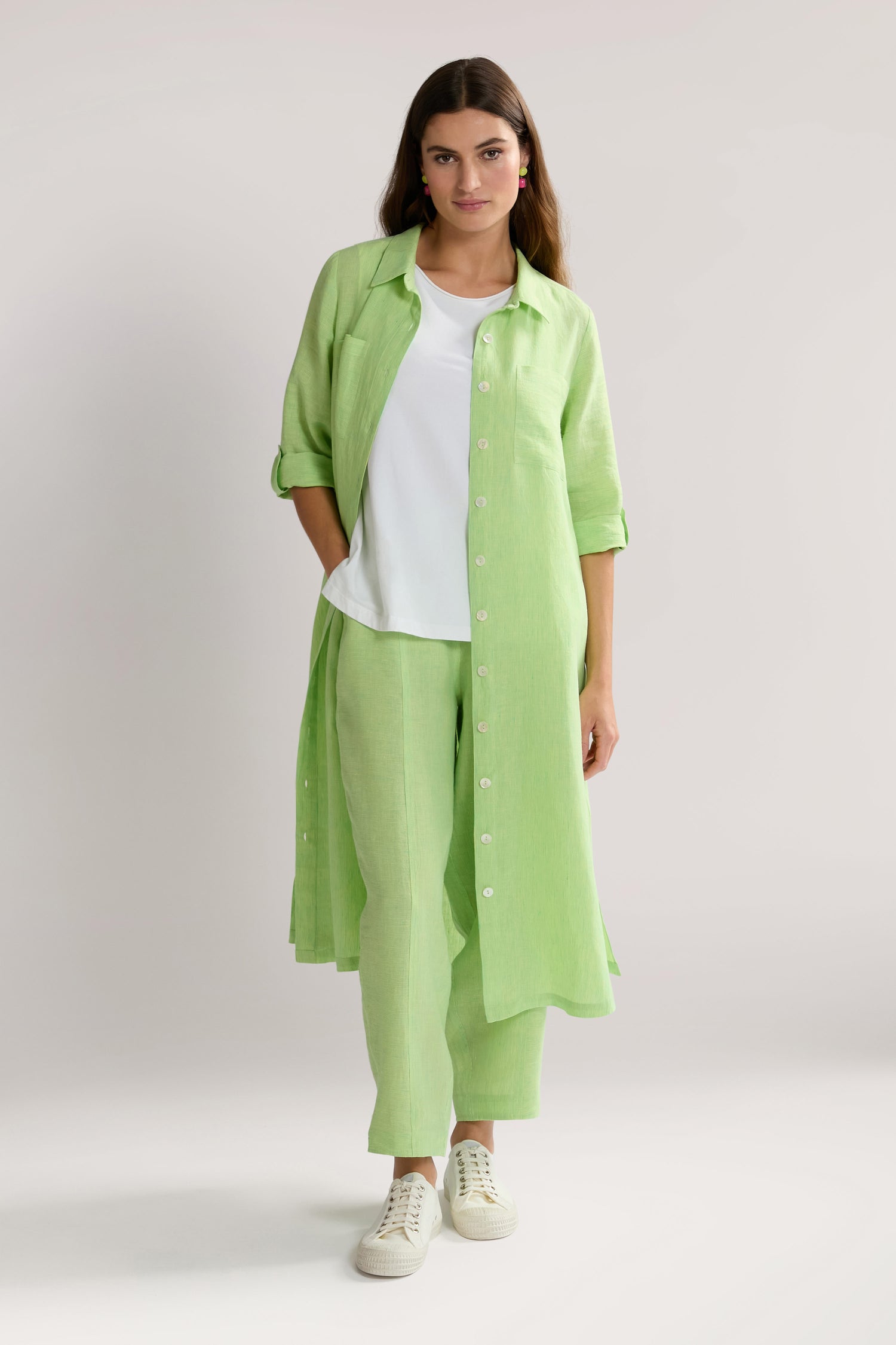 A woman stands against a neutral background wearing a Linen Ticking Stripe Shirt Dress over a white top and matching green pants, paired with white sneakers.