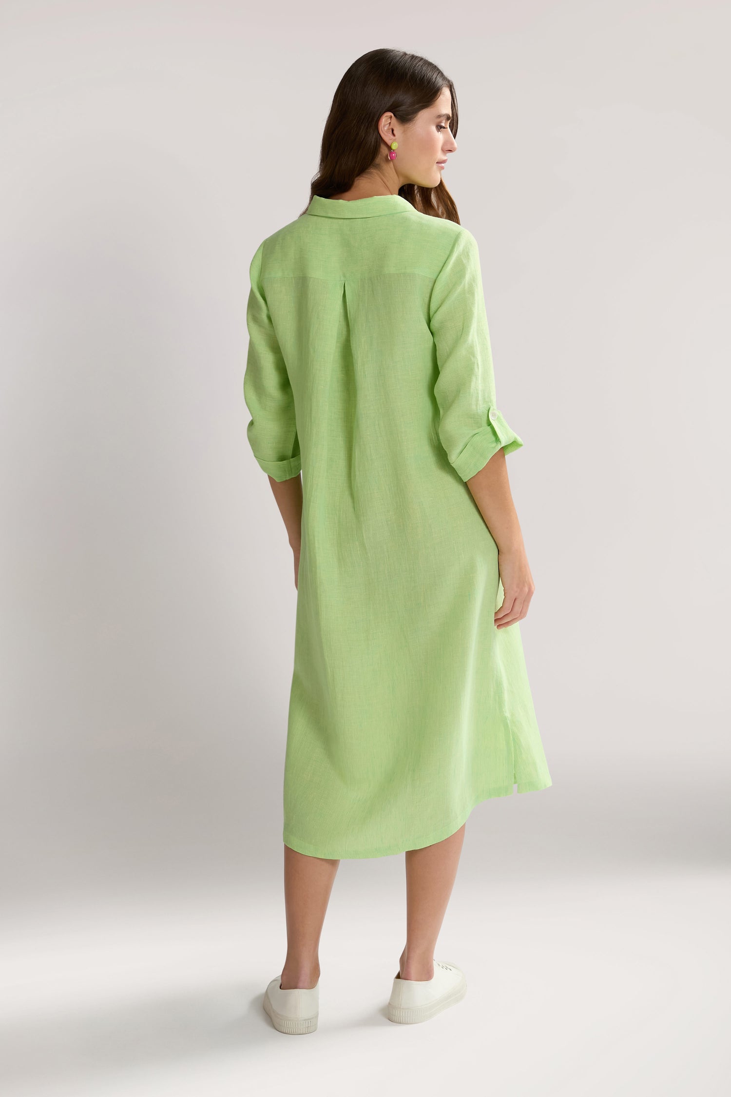 A woman stands facing away, wearing a light green, long-sleeved Linen Ticking Stripe Shirt Dress with rolled-up cuffs and white sneakers. The airy design is complemented by the use of natural fibres, set against a plain, light-colored background.