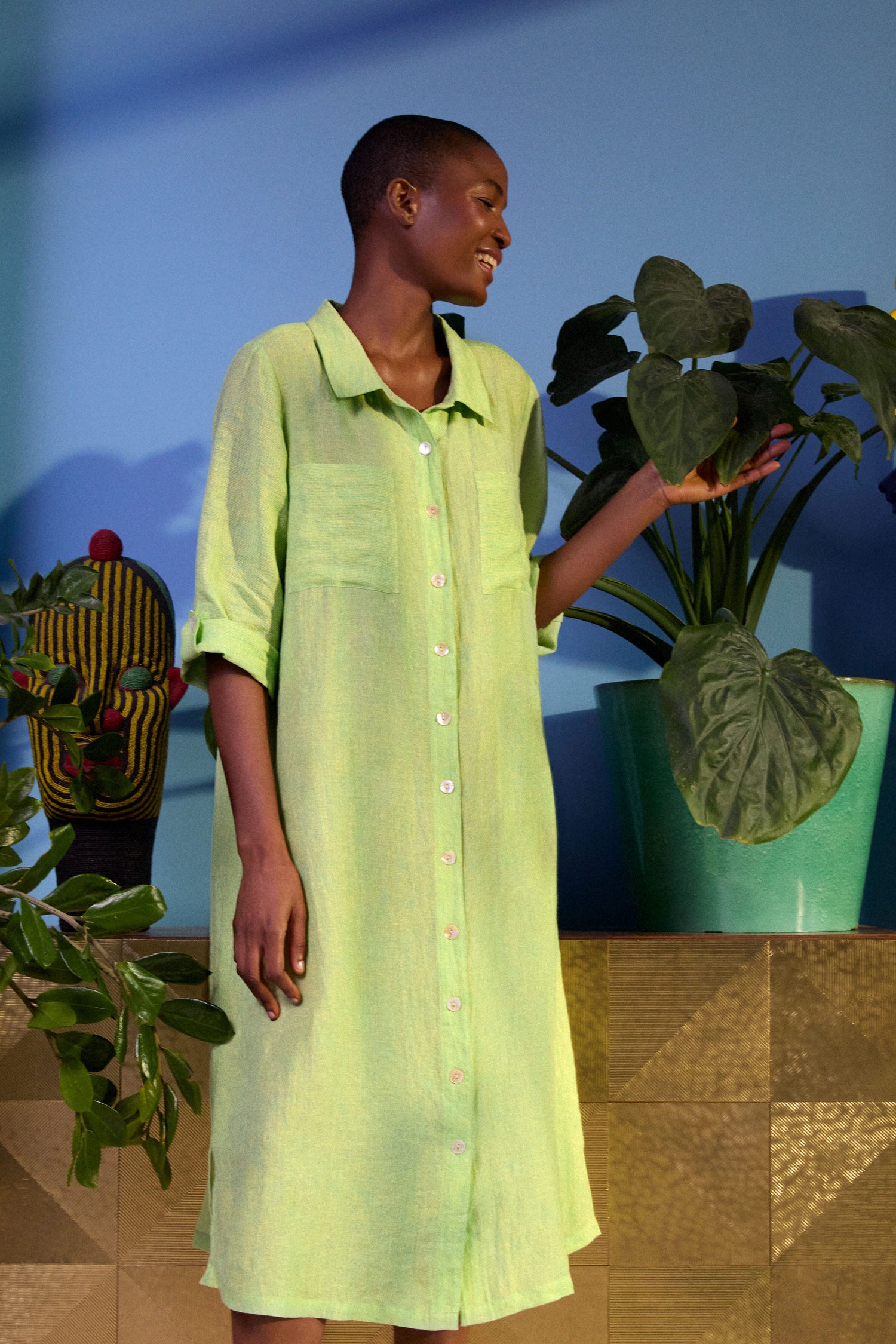 A person stands indoors, gently touching a leaf of a potted plant, wearing a light green Linen Ticking Stripe Shirt Dress made from natural fibers.