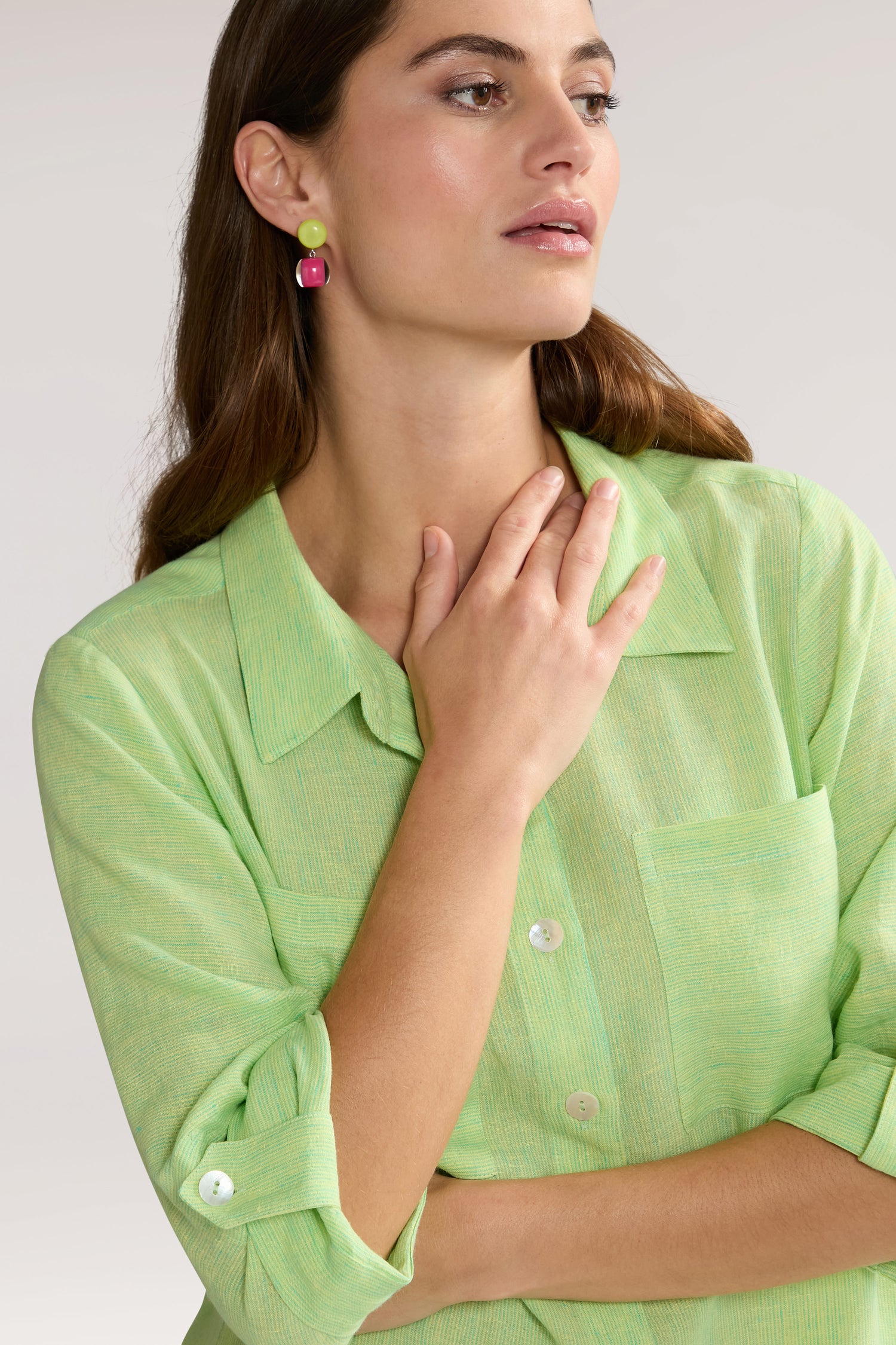 A woman in a light green Linen Ticking Stripe Shirt Dress made from natural fibres with rolled-up sleeves touches her chest. She wears pink and green earrings and has long brown hair, accentuating the airy design of her attire.