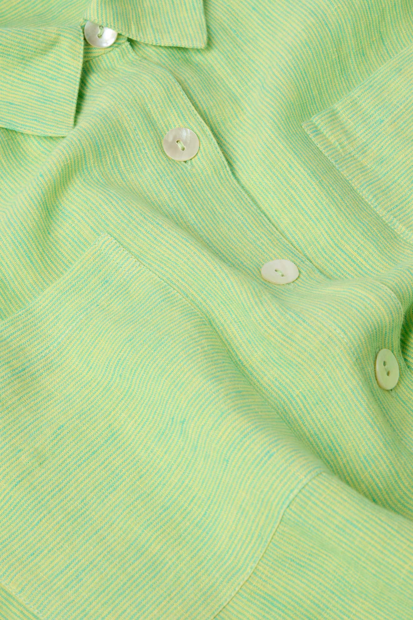 Close-up of a light green Linen Ticking Stripe Shirt Dress made from natural fibres with a soft, textured fabric. The relaxed style shirt dress features a pocket and four white buttons visible, emphasizing its casual elegance.