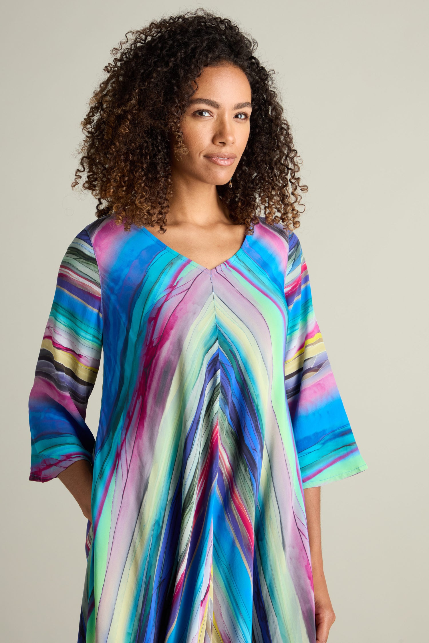 A person with curly hair wears the vibrant Watercolour Landscape Bubble Dress, featuring a V-neckline and 3/4 sleeves, enhanced by its flattering silhouette.