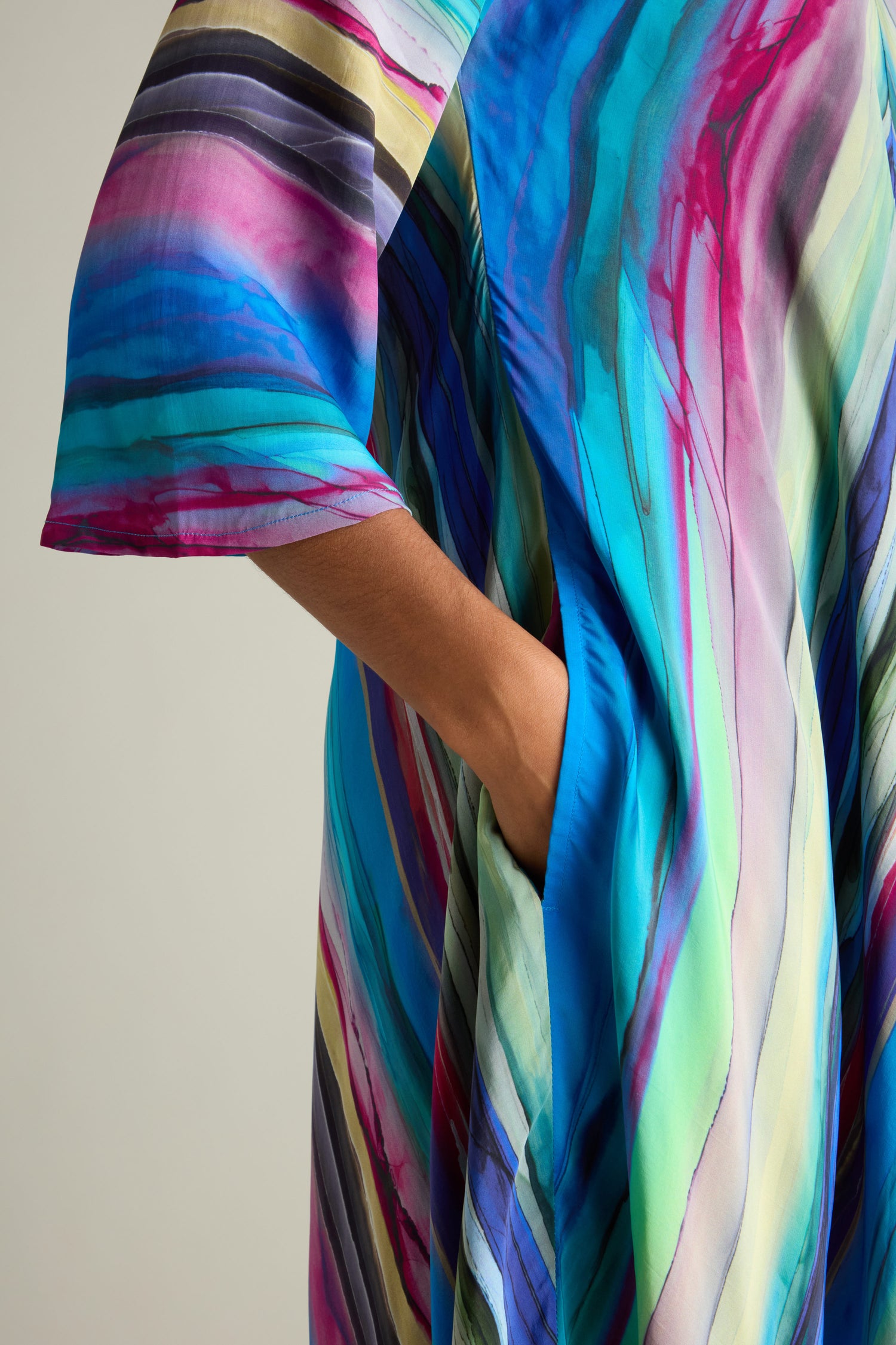 A close-up of a person wearing the Watercolour Landscape Bubble Dress, a colorful garment with shades of blue, pink, and green. The lightweight viscose fabric creates a flattering silhouette as the person casually rests their hand in one of its pockets.