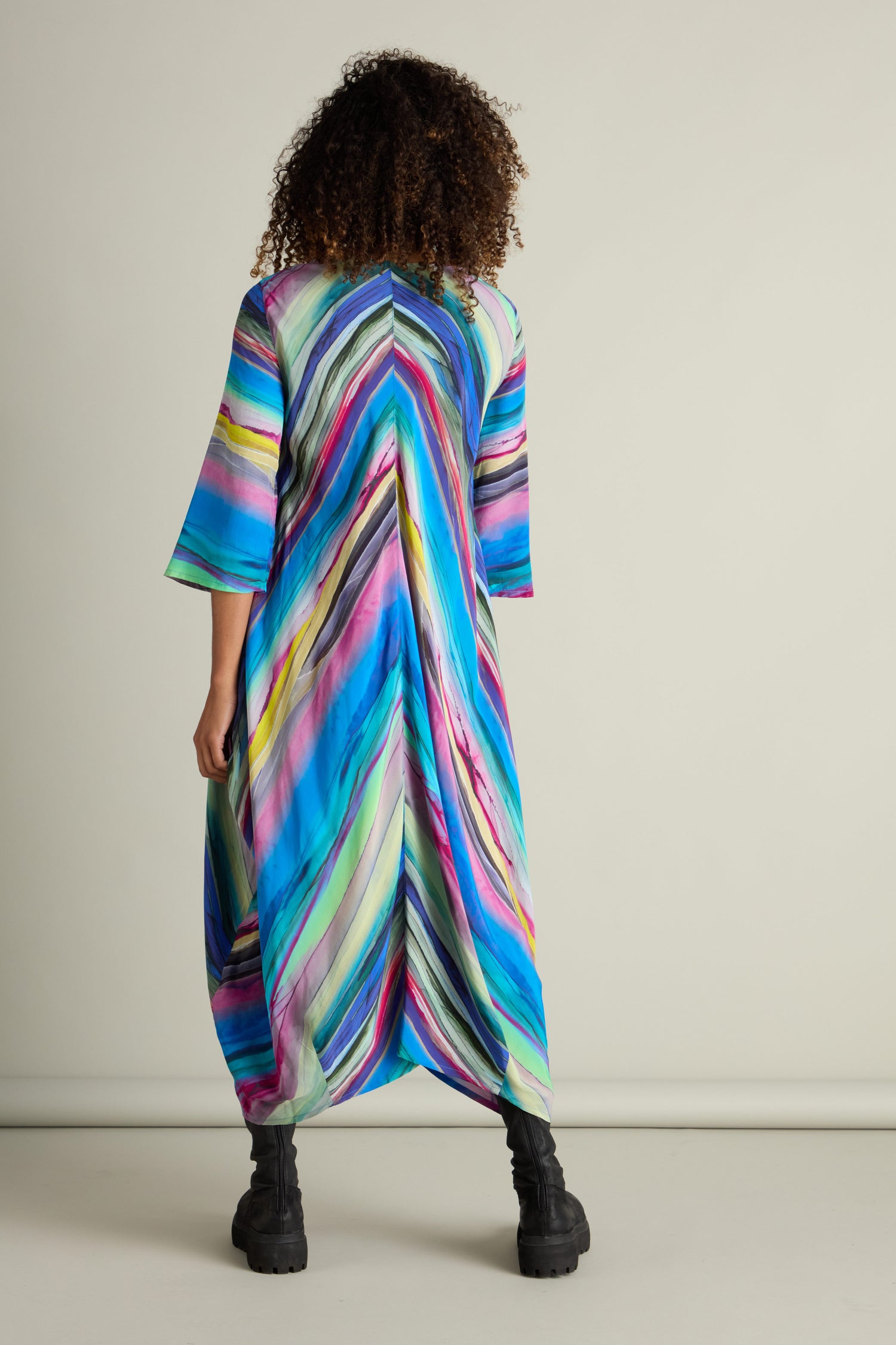 A person with curly hair stands facing away, wearing the Watercolour Landscape Bubble Dress that showcases a vibrant chevron pattern on its lightweight viscose fabric.