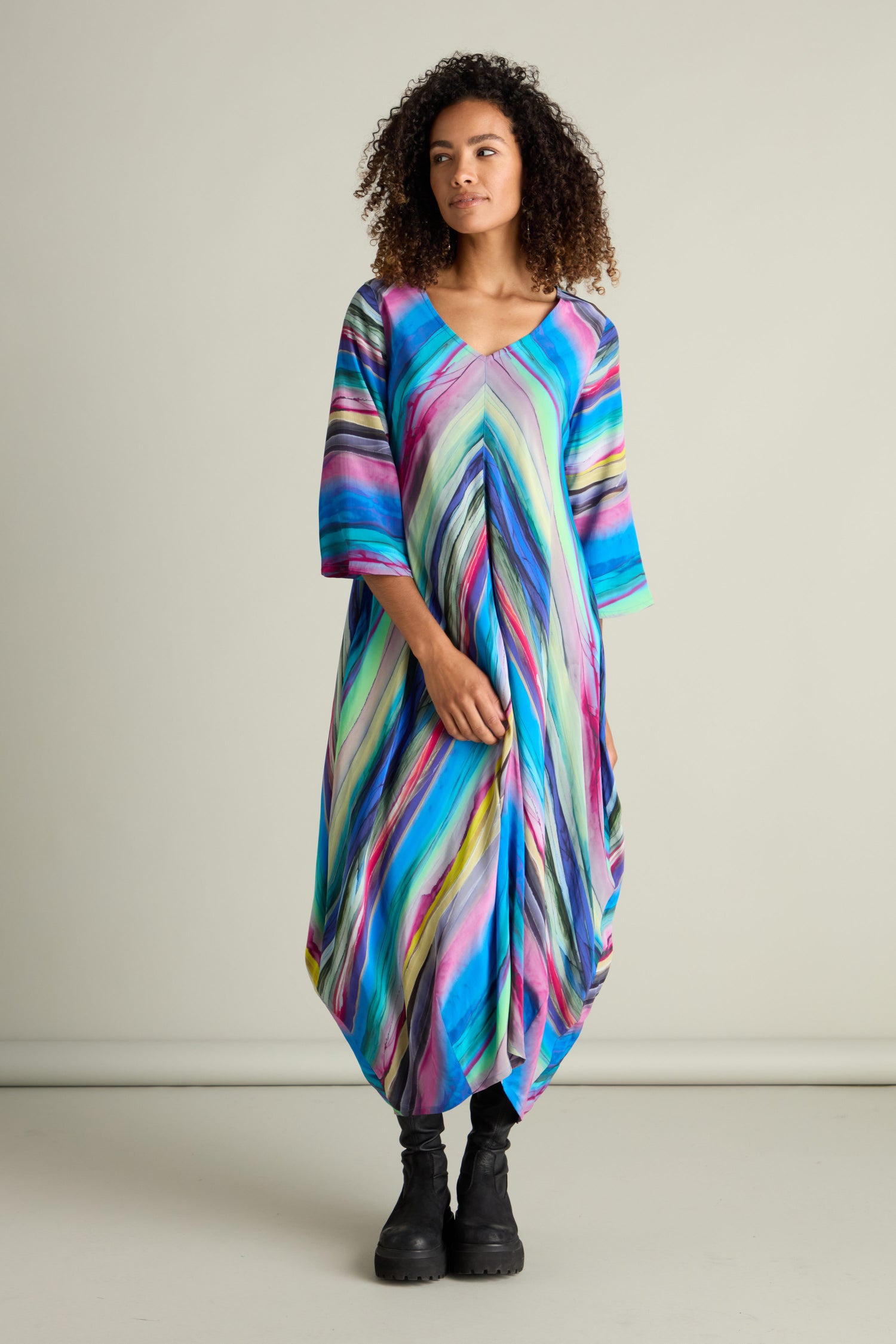 A person stands wearing a colorful, striped Watercolour Landscape Bubble Dress, featuring a v-neck and half sleeves, made from lightweight viscose fabric. They have paired it with black boots. The background is plain and light-colored.