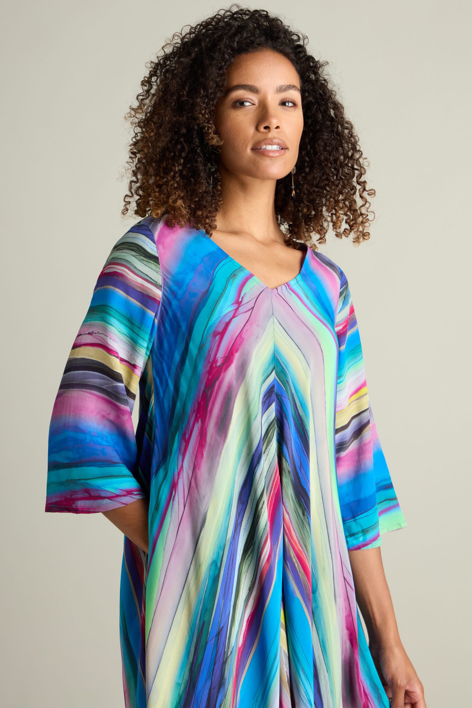 A person with curly hair is wearing the Watercolour Landscape Bubble Dress, a colorful, striped creation crafted from lightweight viscose fabric in shades of blue, pink, and green. They are standing against a plain background.