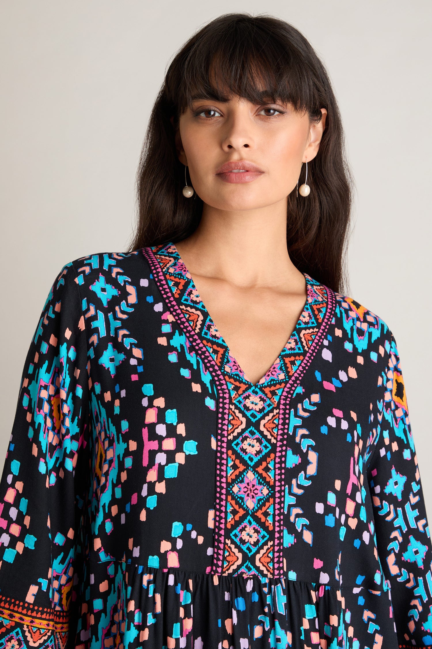 A person with straight dark hair, dressed in the vibrant Geo Kilim Dress with its colorful geometric patterns, poses against a neutral background.