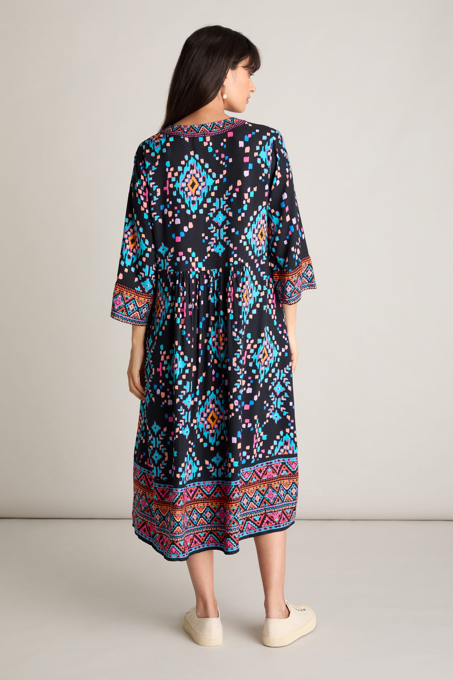 A person stands facing away, showcasing the vivid Geo Kilim Dress. This long A-line silhouette flaunts colorful geometric patterns, with 3/4 sleeves and a hem beautifully adorned with intricate designs in shades of blue, pink, and orange.