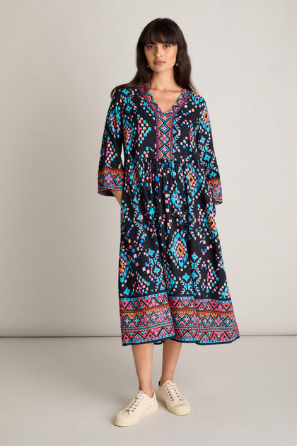 A woman wearing a Geo Kilim Dress featuring long sleeves and an ankle-length cut, adorned with a colorful geometric pattern, stands against a plain background. She has long dark hair with bangs and pairs the dress with white sneakers.